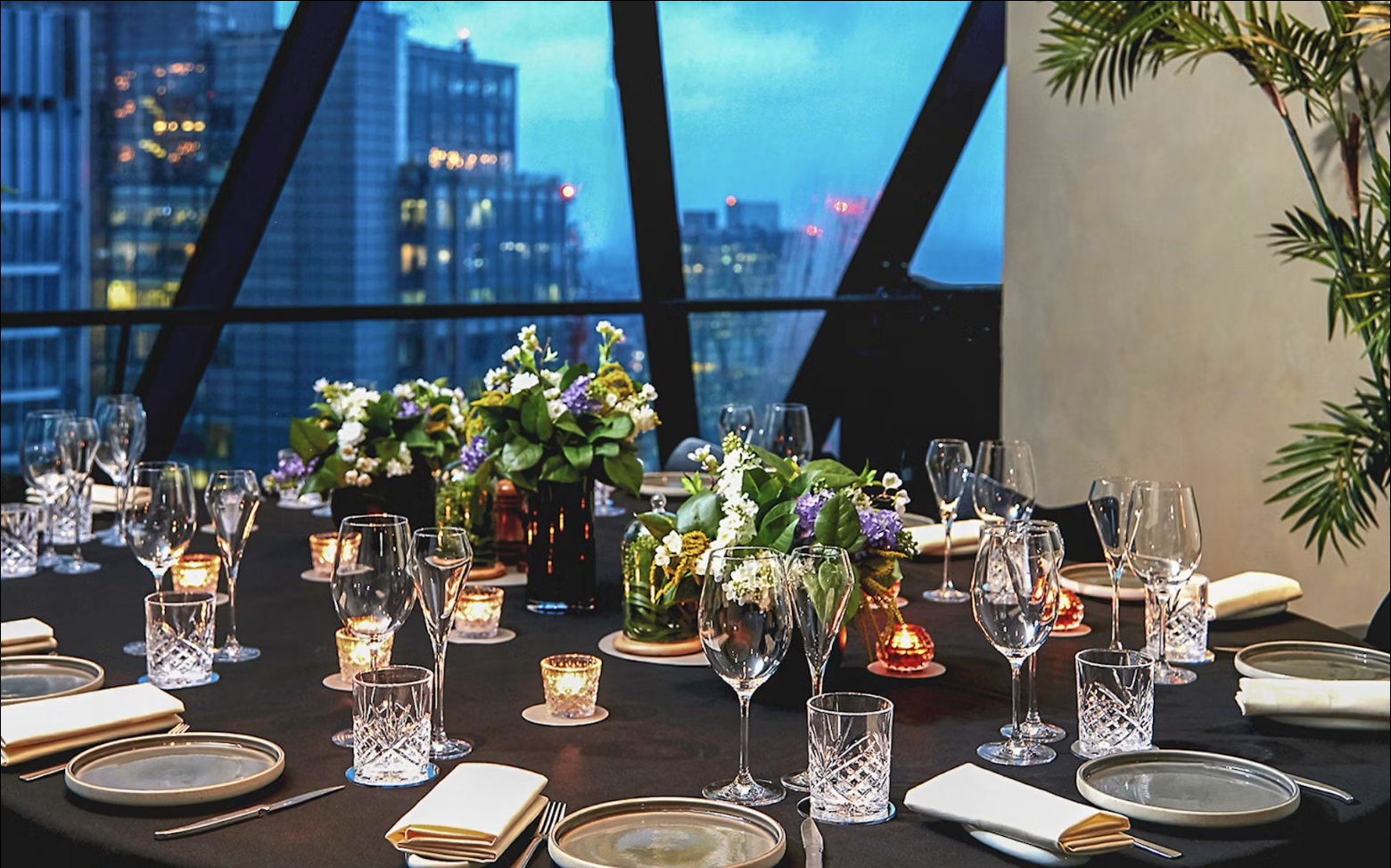 Elegant dining table set for a formal event with floral centerpieces, candles, wine glasses, and napkins, overlooking a cityscape through large windows.