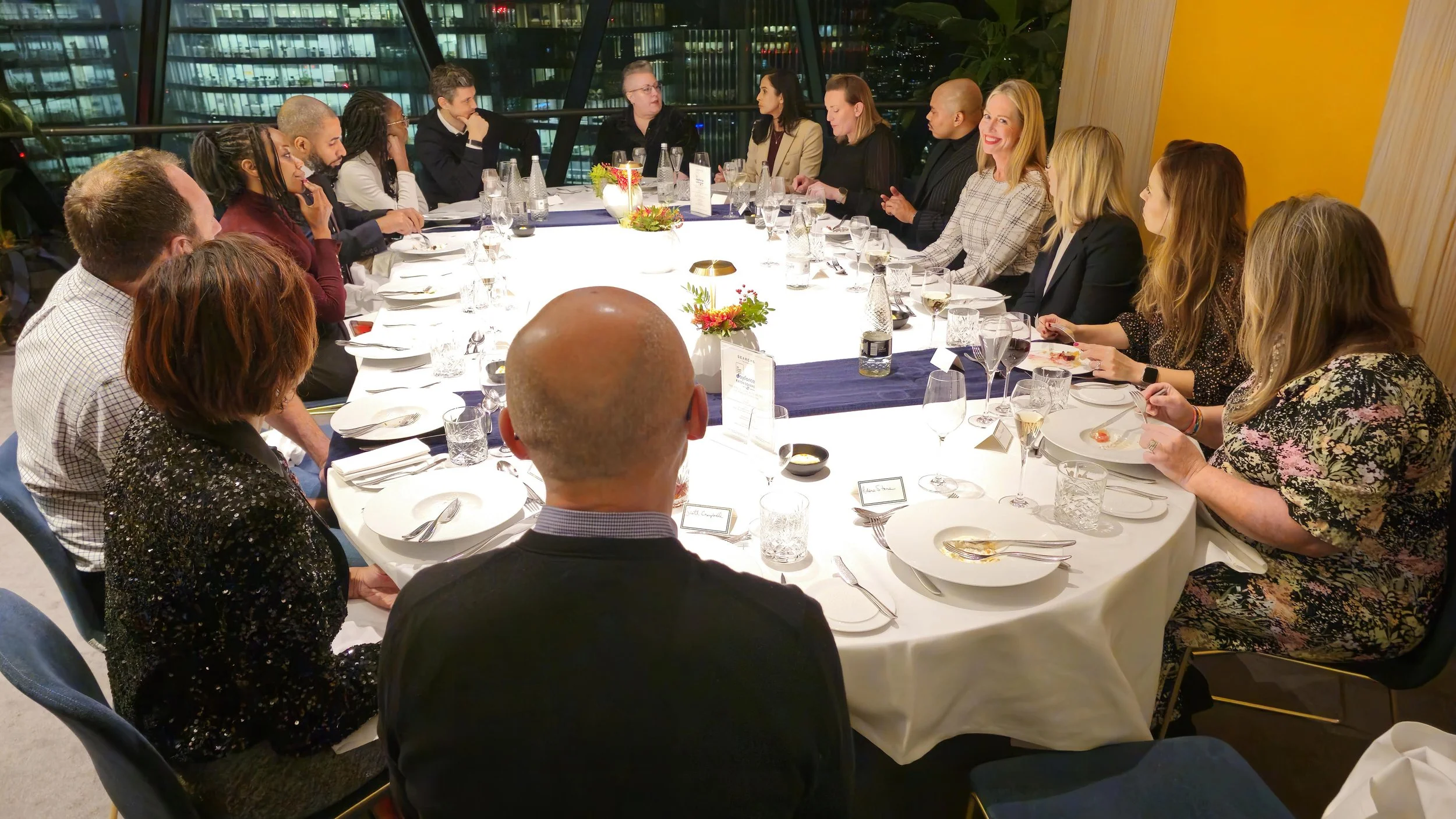 A group of people sitting around a large, round, illuminated dining table in a modern high rise building at night, engaged in conversation.