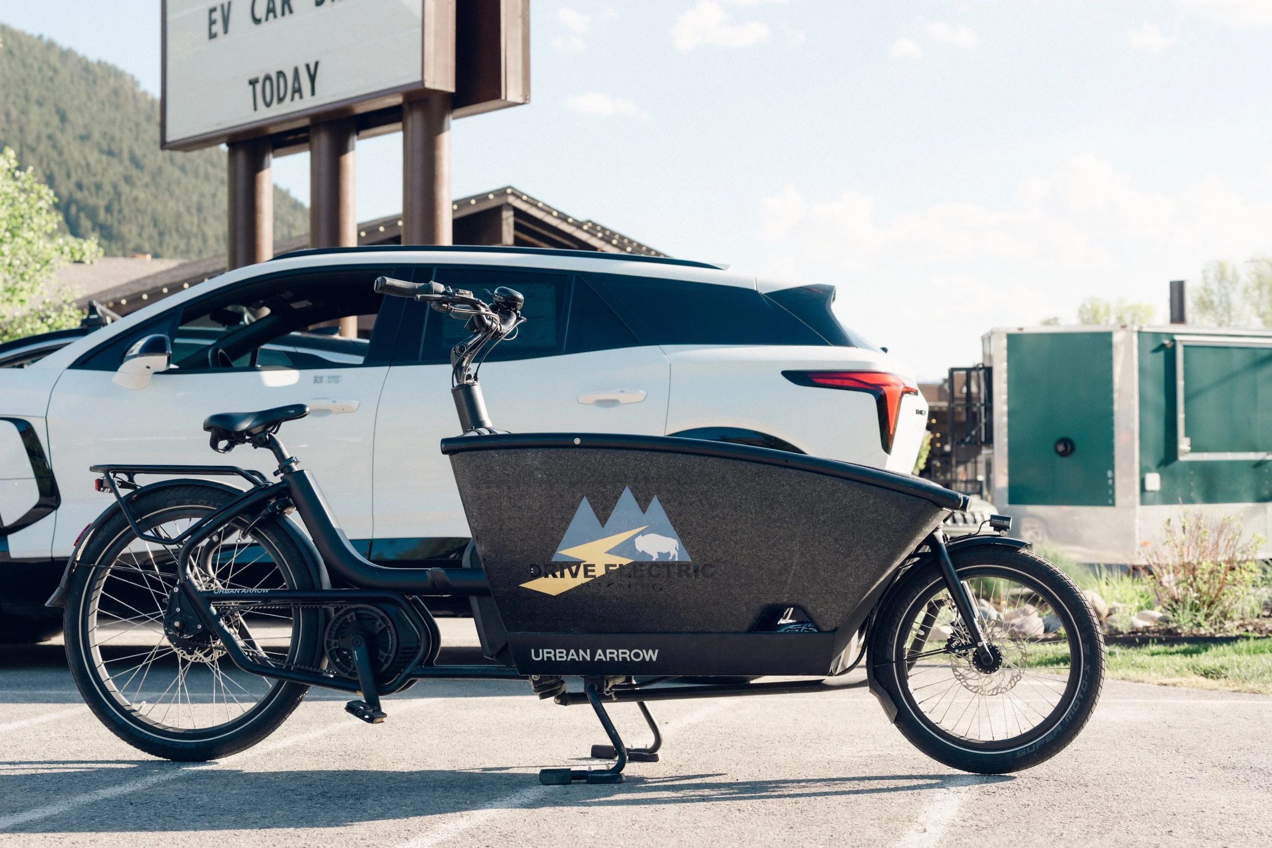 Electric delivery bike parked in front of a white car at a parking lot, with a sign above and a mountain in the background.