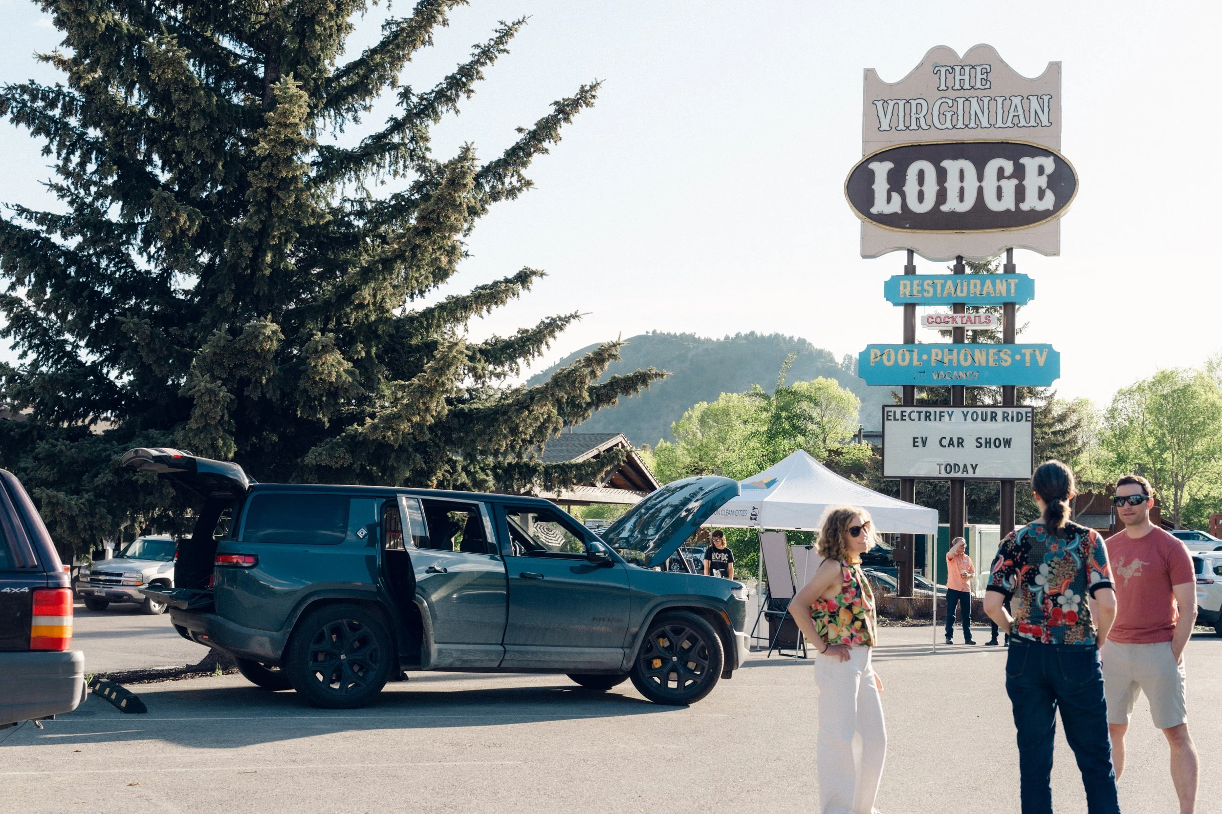 People standing near a vintage car with an open hood in a parking lot, with a large sign for The Virginian Lodge in the background.