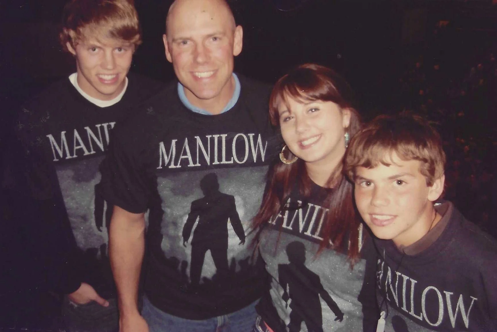 Family of four wearing matching 'Manilow' t-shirts, posing together outdoors at night, smiling at the camera.