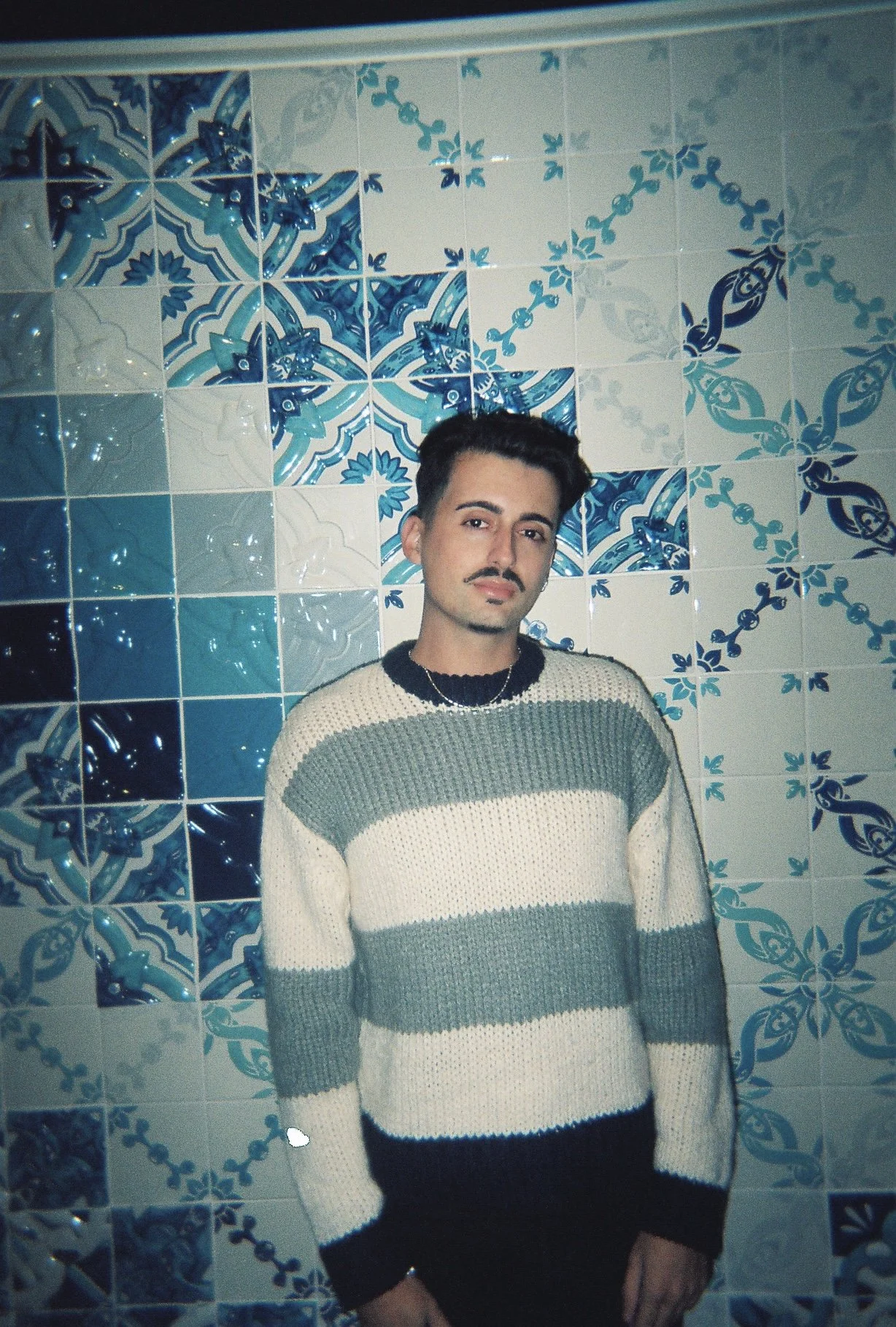 Young man with dark hair and a mustache standing in front of a blue and white decorative tiled wall, wearing a striped sweater.