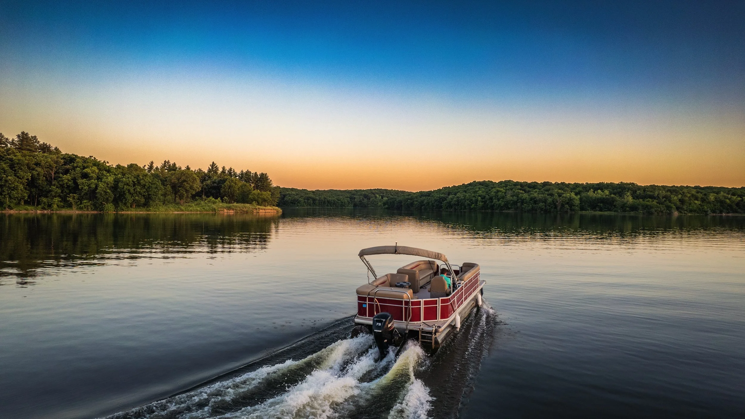 A pontoon boat sailing on a calm lake during sunset with green trees along the shoreline.