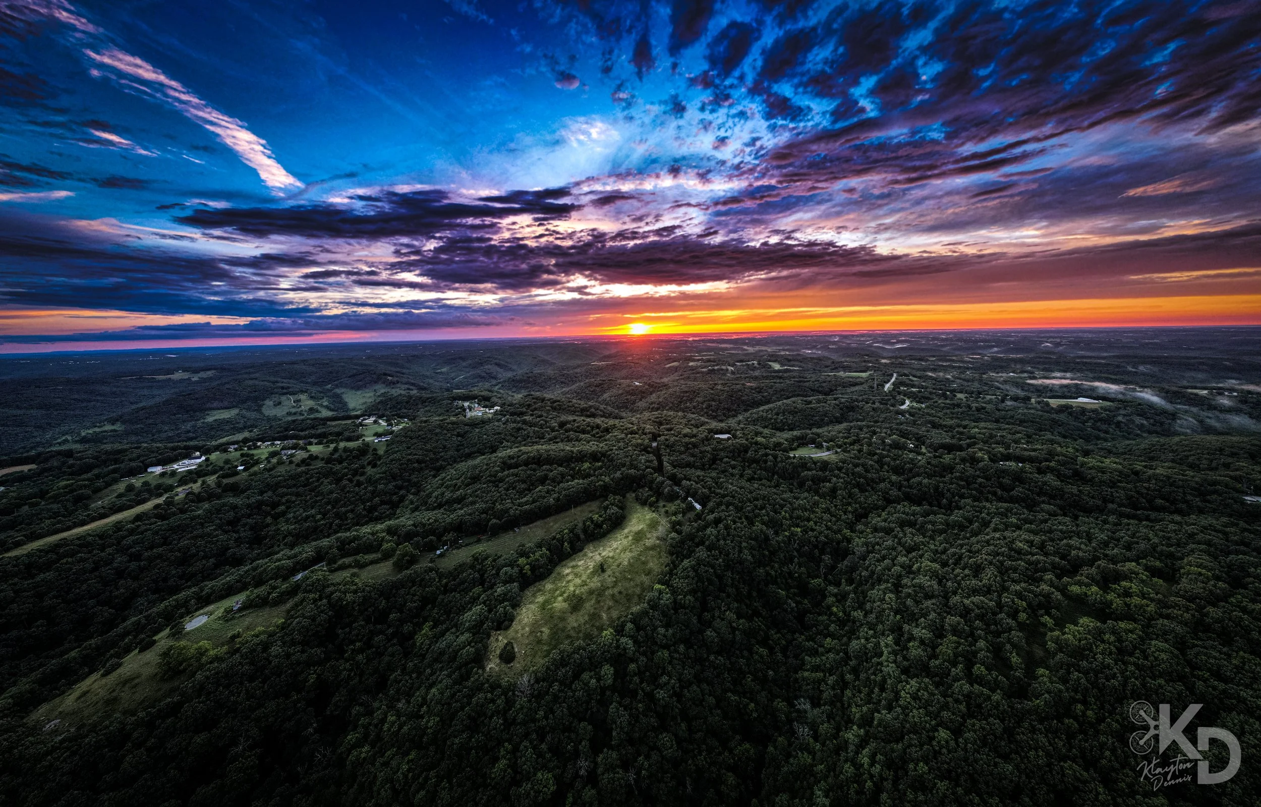 Aerial view of a vast landscape with rolling green hills, scattered trees, and a sunset with dramatic colorful clouds in the sky.