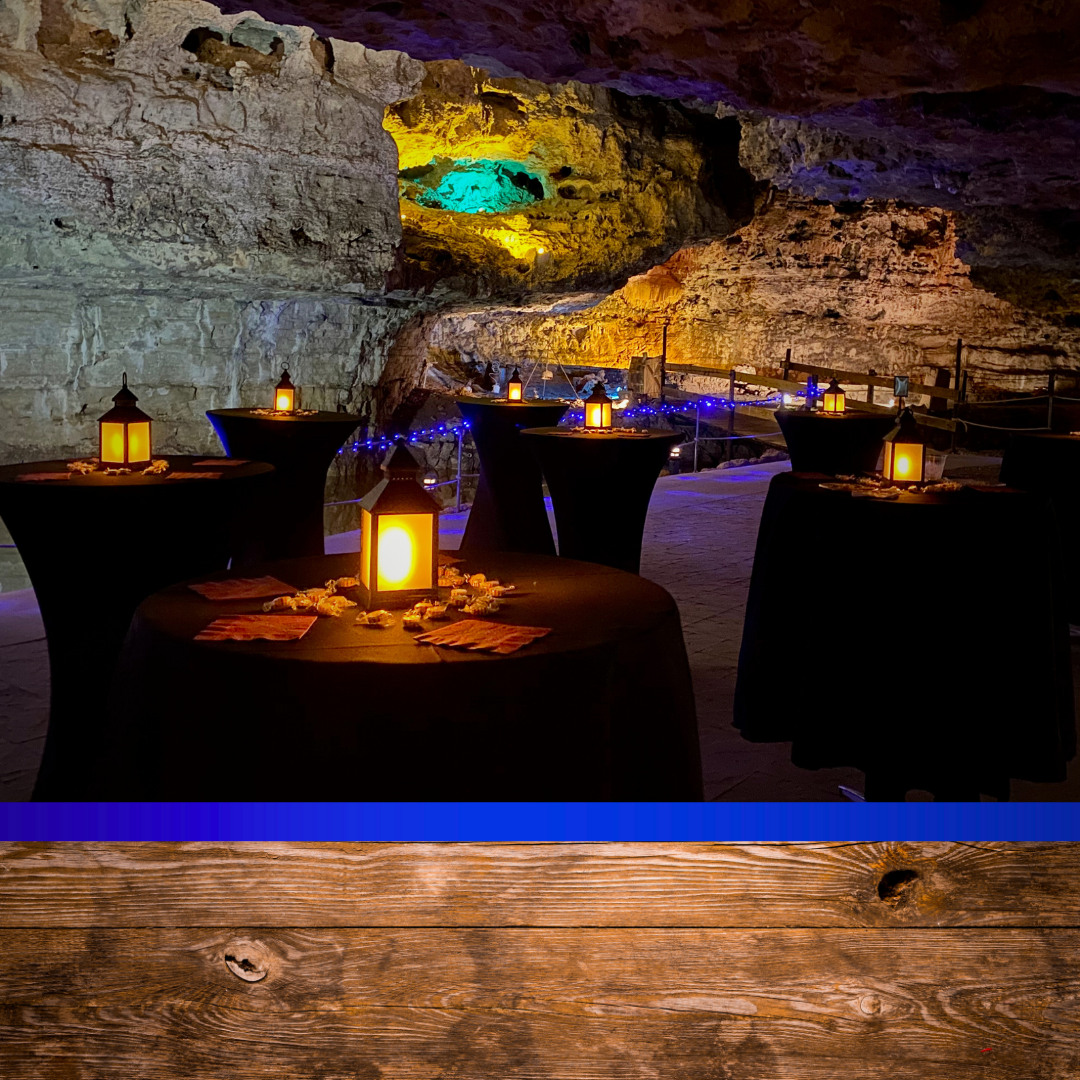 Underground cave decorated with tables covered in black cloths, each with lanterns and table settings. Cave walls and ceiling illuminated with colorful lights, creating a cozy atmosphere for an event.