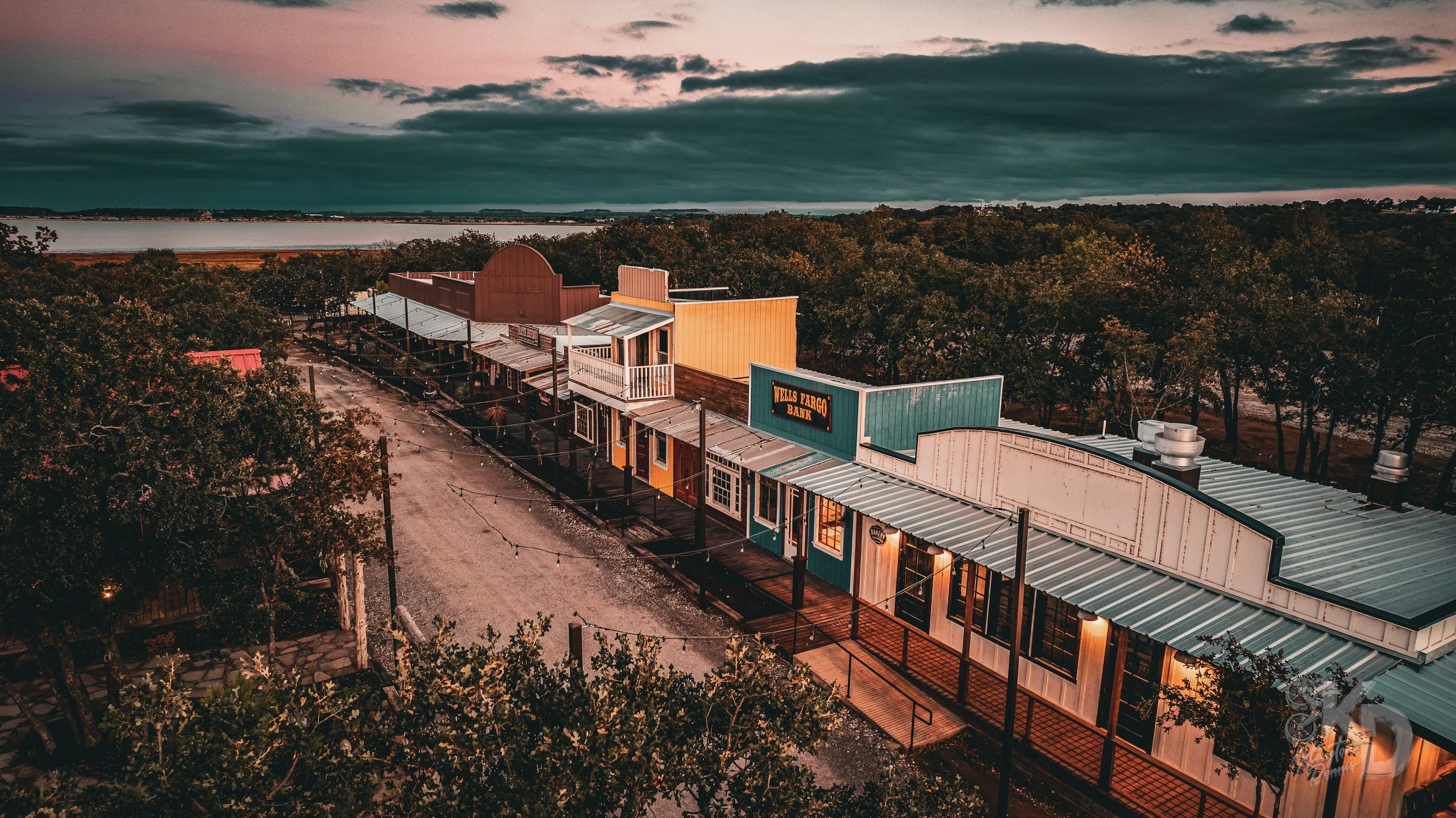 Aerial view of a rustic small town street at sunset with colorful storefronts and a 'Wells Fargo Bank' sign, surrounded by trees and a large body of water in the background under a cloudy sky.