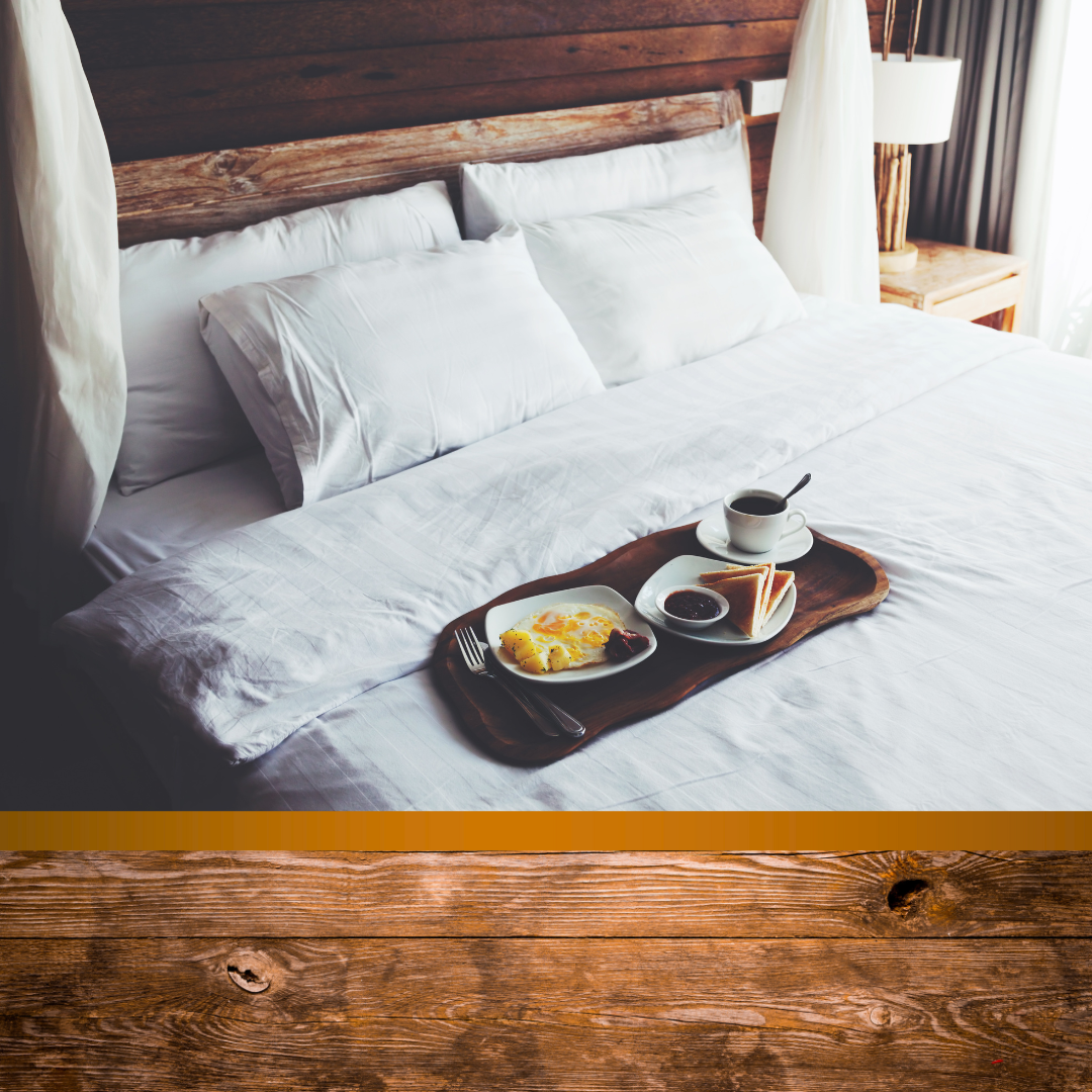 Breakfast tray with fried eggs, toast, jam, and a cup of coffee on a wooden bed tray in a cozy bedroom with white bedding and a wooden headboard.