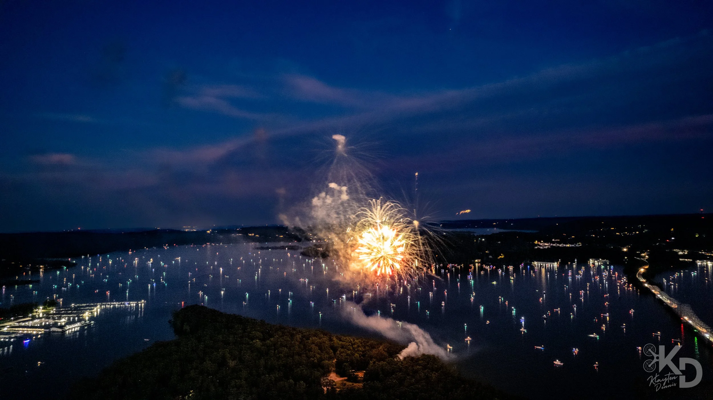 Nighttime scene of fireworks lighting up the sky over a large body of water, with boats and docked ships reflecting the fireworks' colors and lights.