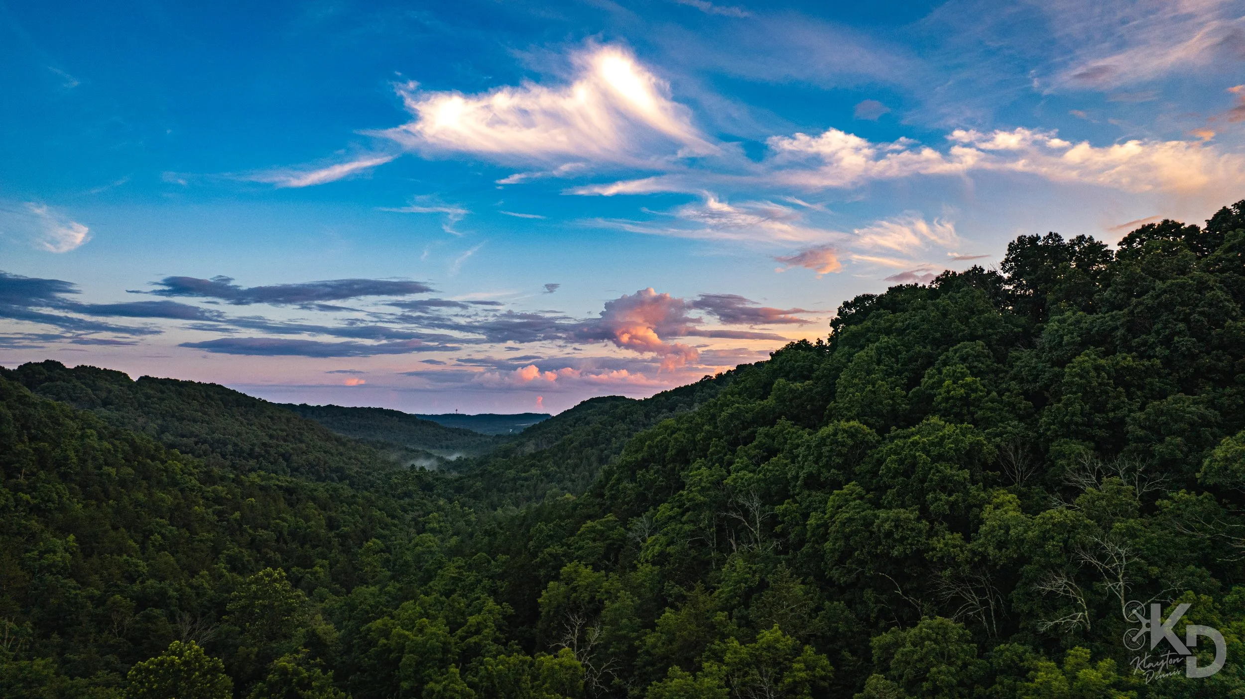 A scenic view of a lush green mountain valley during sunset with a sky filled with wispy clouds and a soft pink glow.
