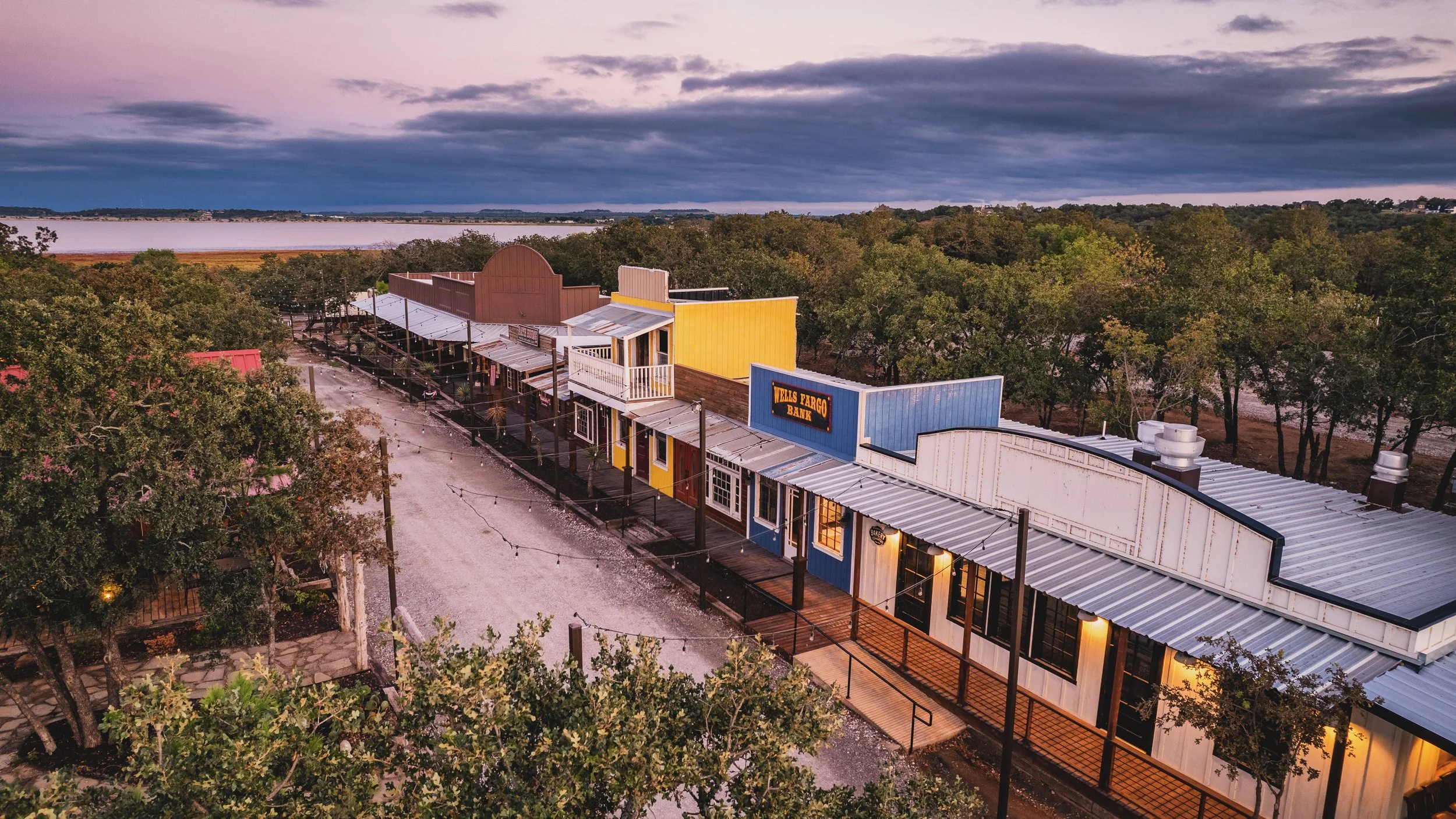 A historic Western-style street with colorful storefronts, including a Wells Fargo Bank sign, at dusk with a cloudy sky and trees in the background.