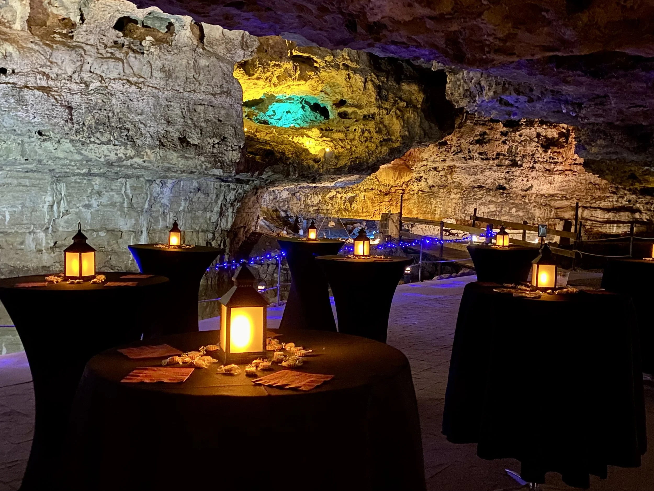 Cave interior with tables covered in black cloth, decorated with lanterns and string lights, illuminated by colorful lighting highlighting the rocky walls and ceiling, creating a cozy atmosphere for an event or gathering.