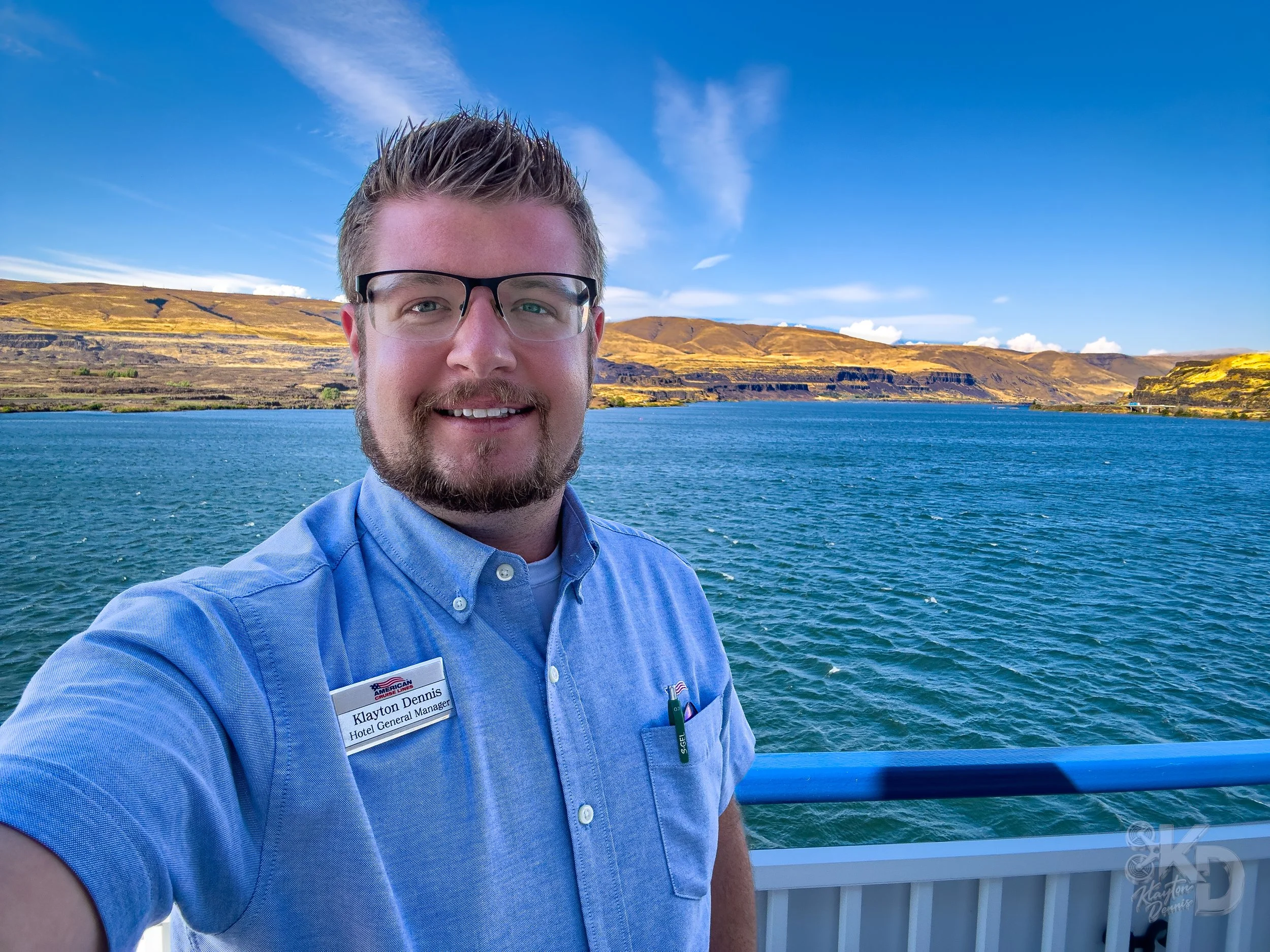 A man with glasses and a light blue shirt smiling for a selfie on a boat, with a lake and rolling hills in the background.