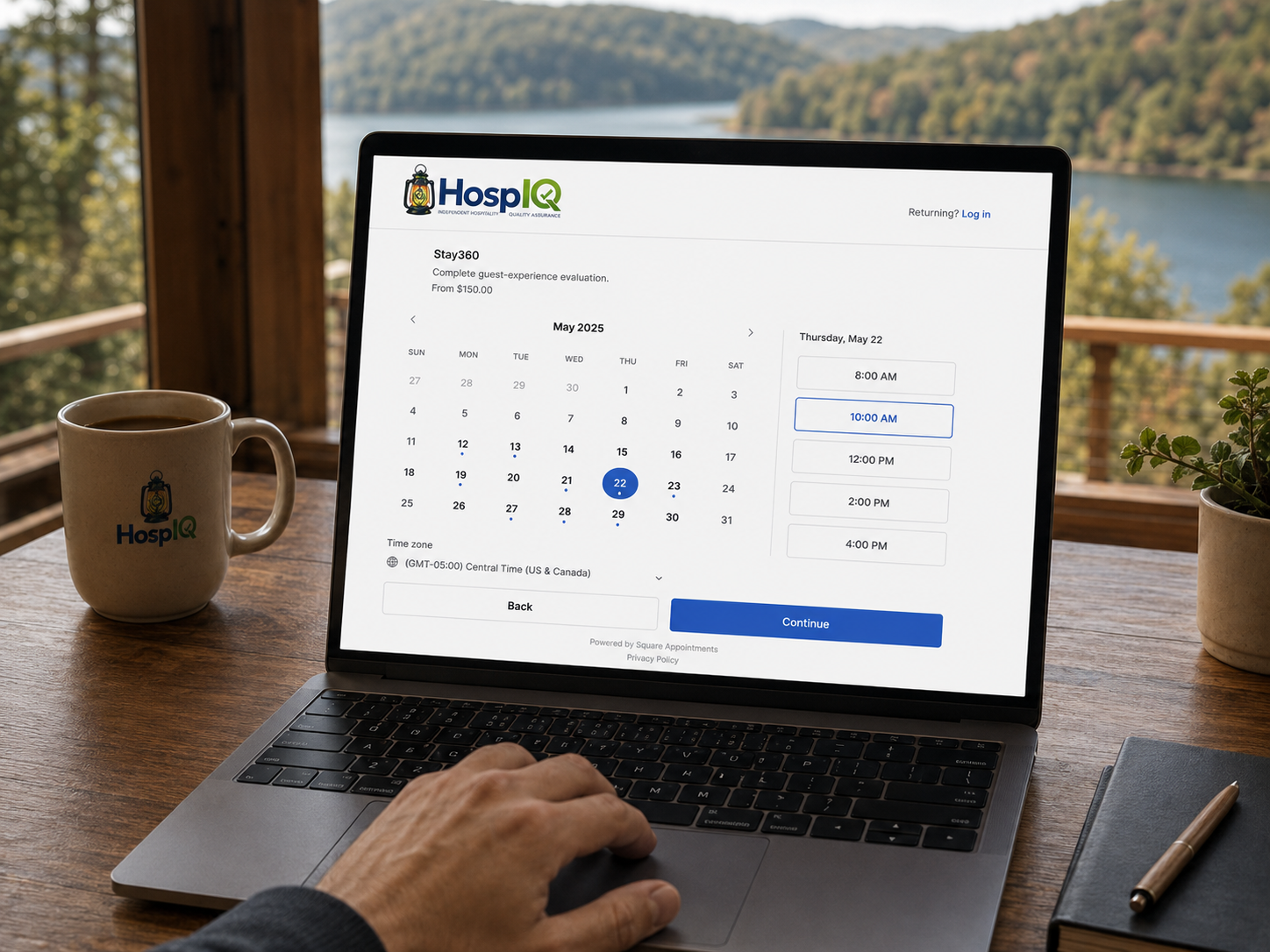 Laptop on a wooden table displaying a scheduling app, with a calendar for May 2025, an option to select in the morning, and a blue continue button. Next to the laptop is a coffee mug with a logo and a potted plant, overlooking a lake and forested hills.