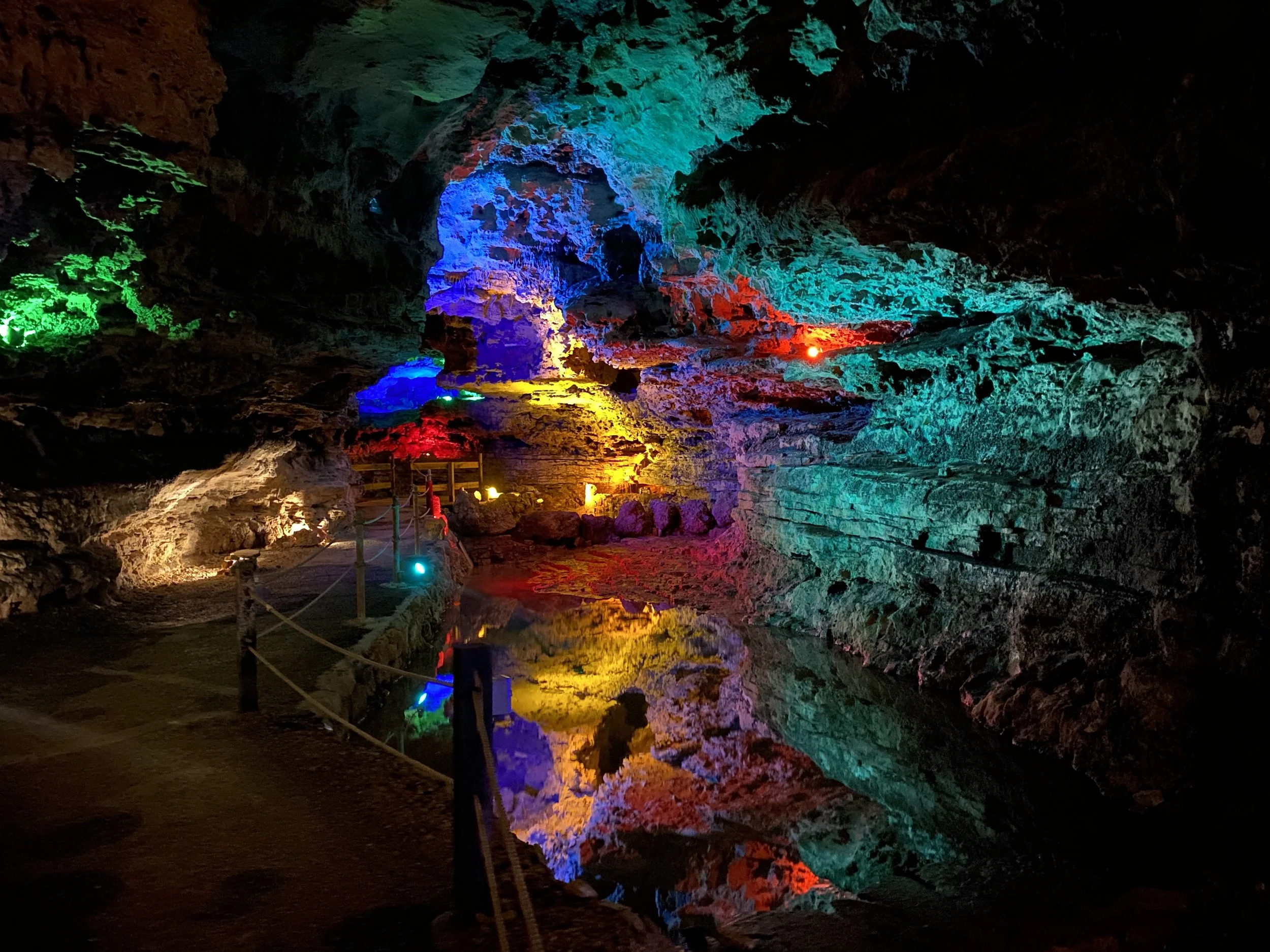 Colorfully lit cave interior with a pathway and a reflective pool, illuminated by multi-colored LED lights highlighting the rock formations.