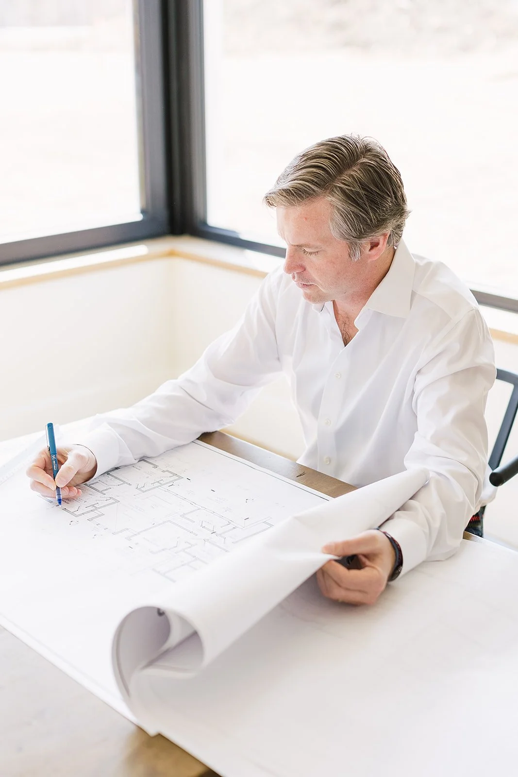 A man in a white shirt reviewing architectural blueprints at a table near large windows.