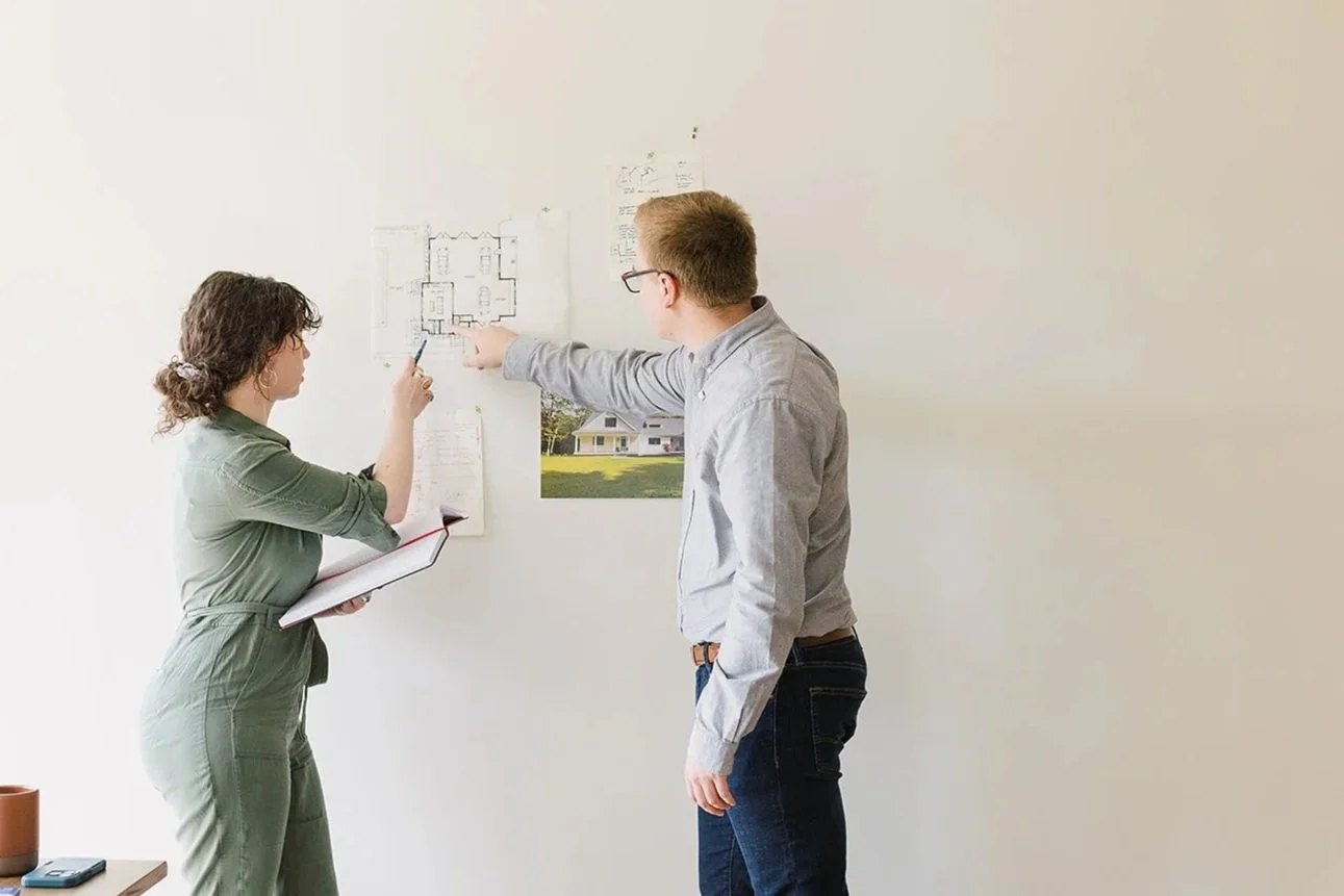 Two people discussing a house plan on a white wall, one pointing to the plans, the other holding a notebook, in a bright room.