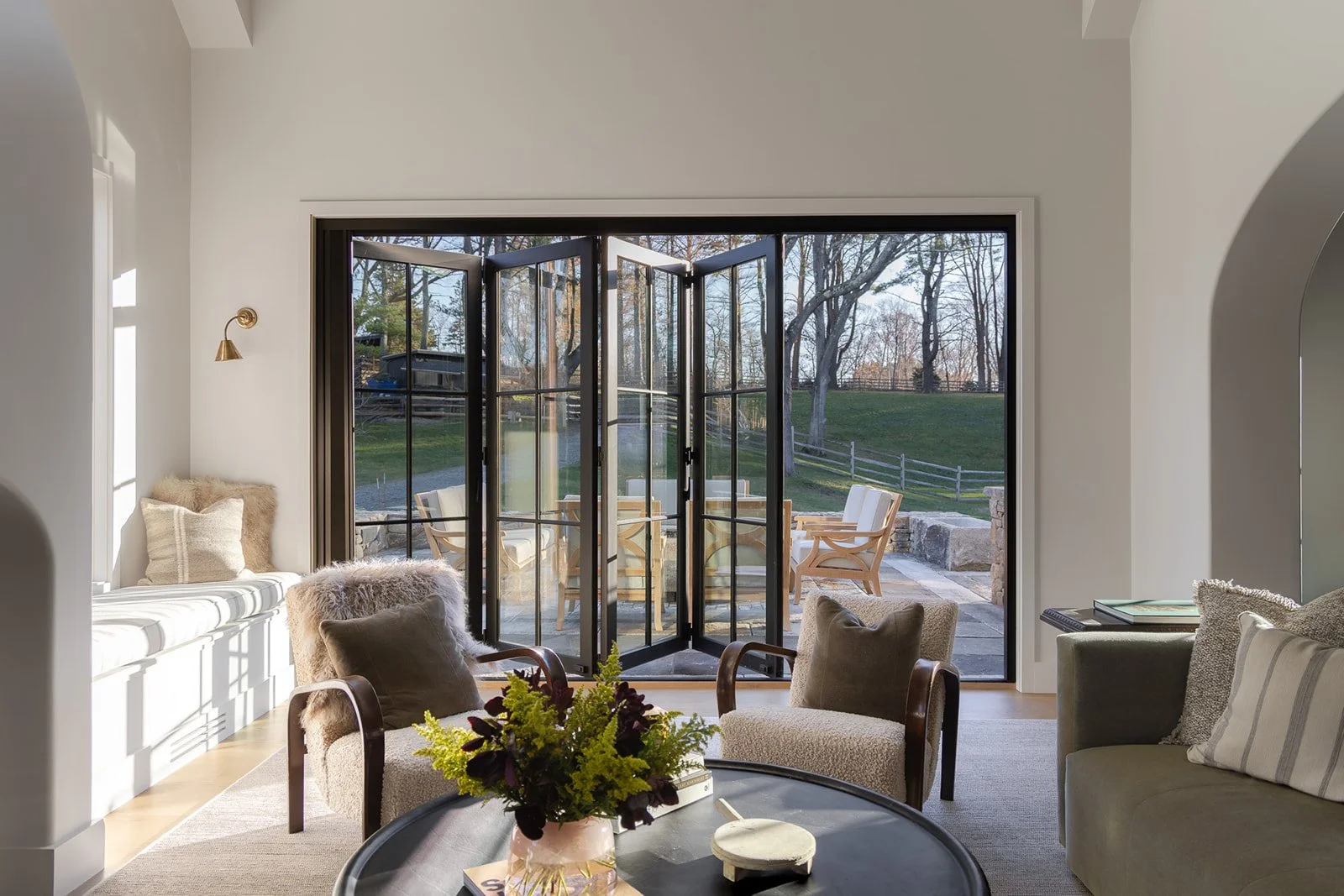 Living room with open glass doors leading to outdoor patio, with outdoor chairs and view of trees and grassy yard.