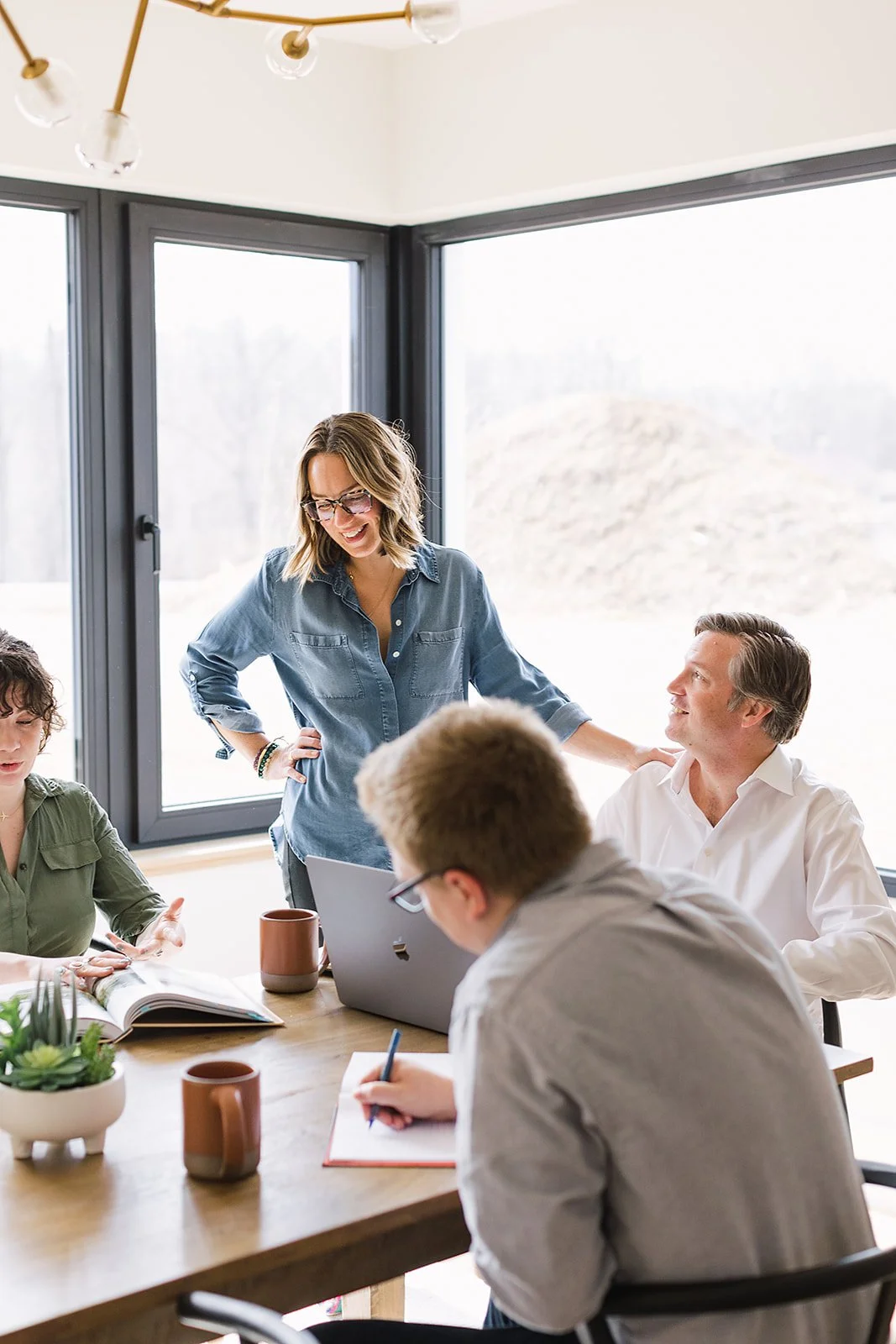 A woman standing and smiling at a conference table with four seated people, near large windows with outdoor scenery. The group is engaged in discussion, with notebooks, mugs, and a laptop on the table.