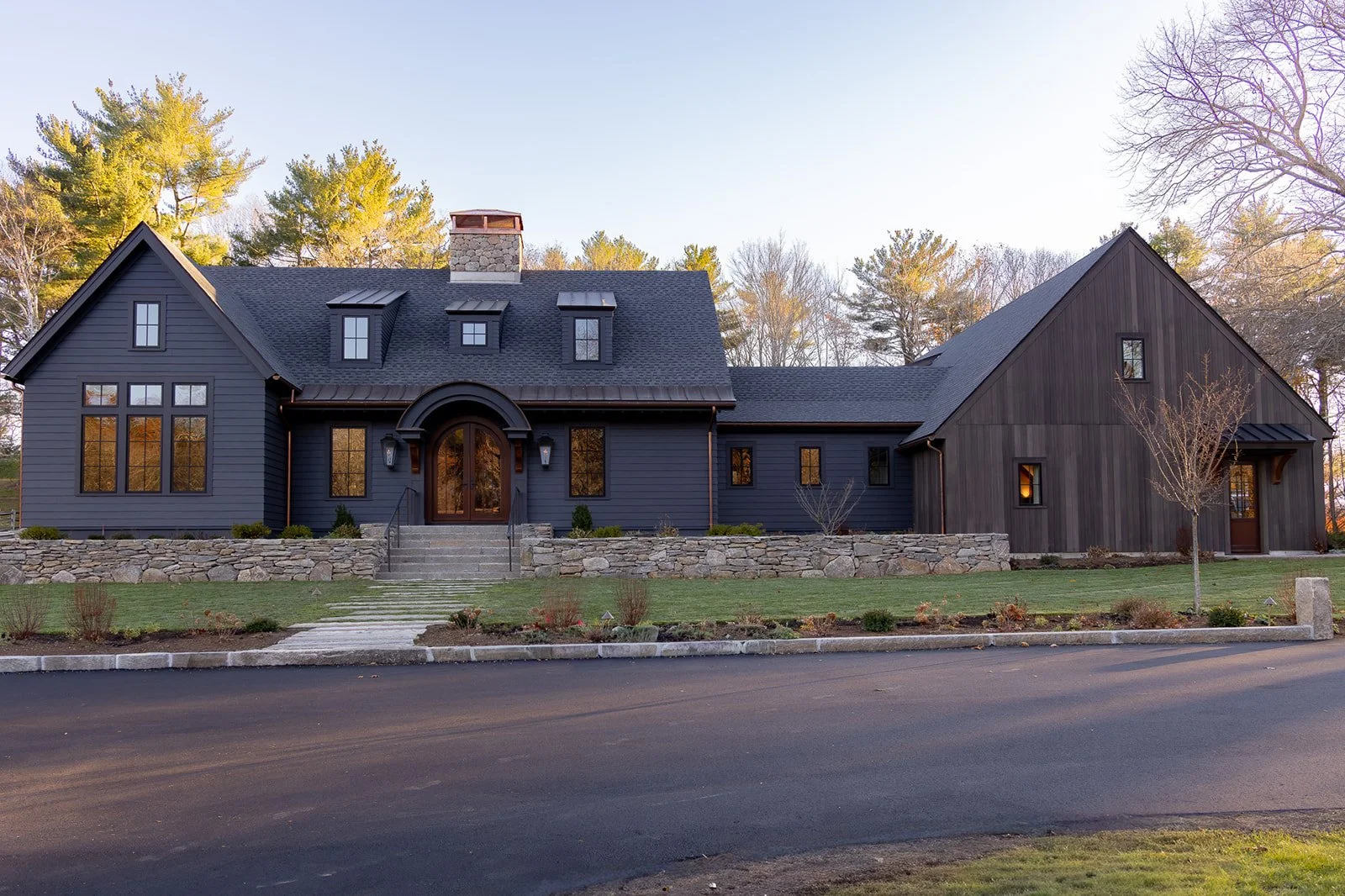 A modern two-story house with dark siding, stone accents, and a steep roof, surrounded by a well-manicured lawn and trees, with a paved road in the foreground.