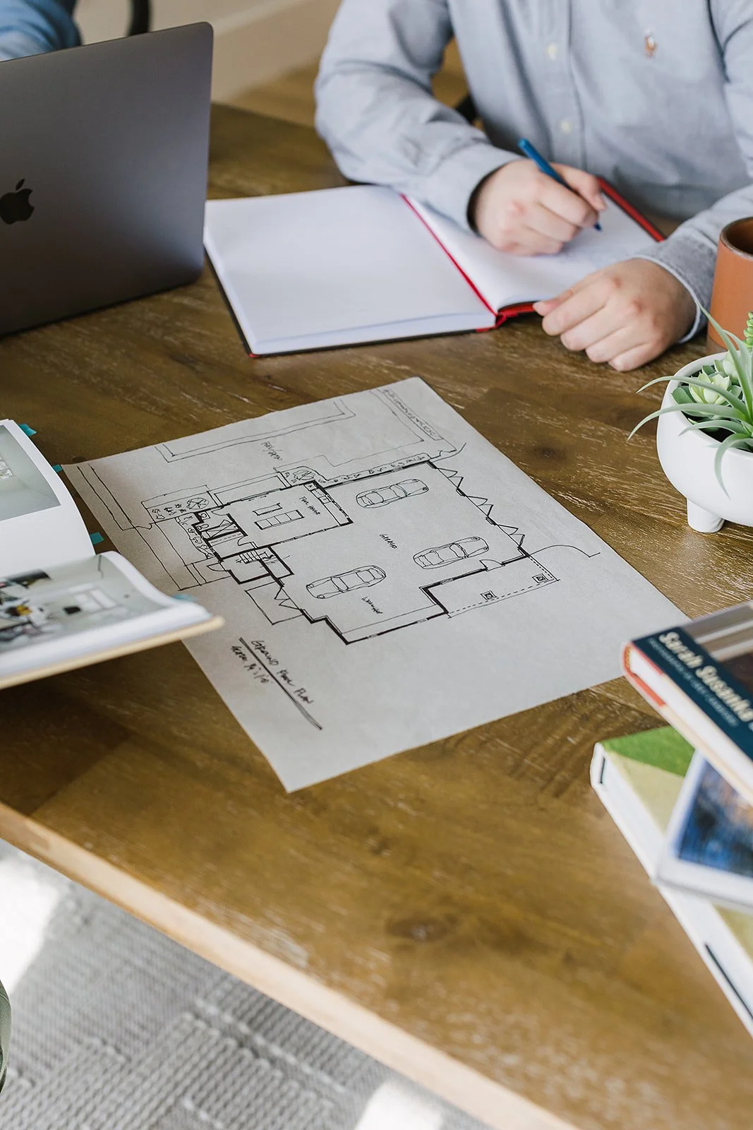A person working on architectural plans for a house, with a laptop, notebooks, and books on a wooden table.