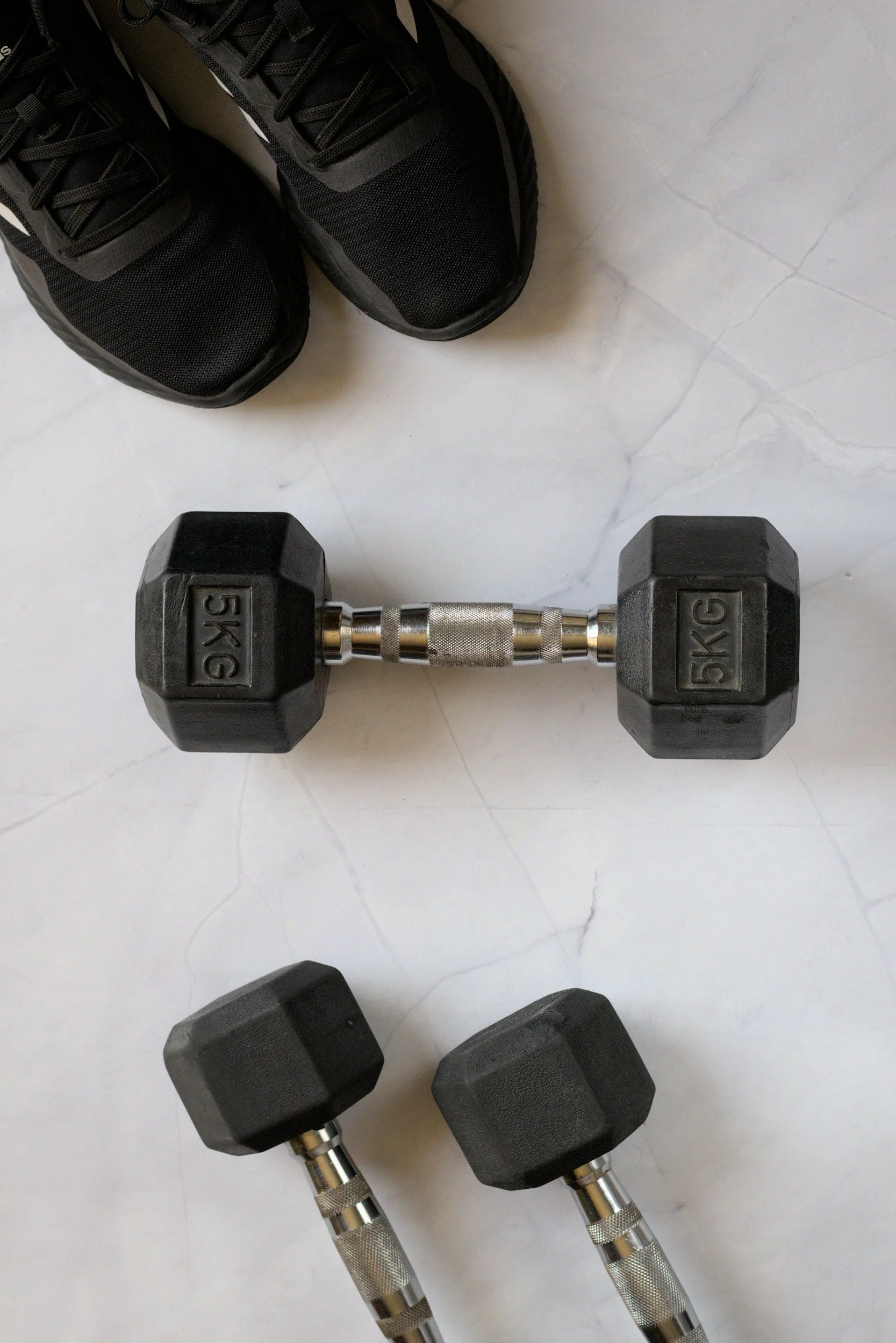 Two pair of black dumbbells, one labeled 5kg and the other with no visible weight, along with a pair of black athletic shoes, are placed on a white marble floor.