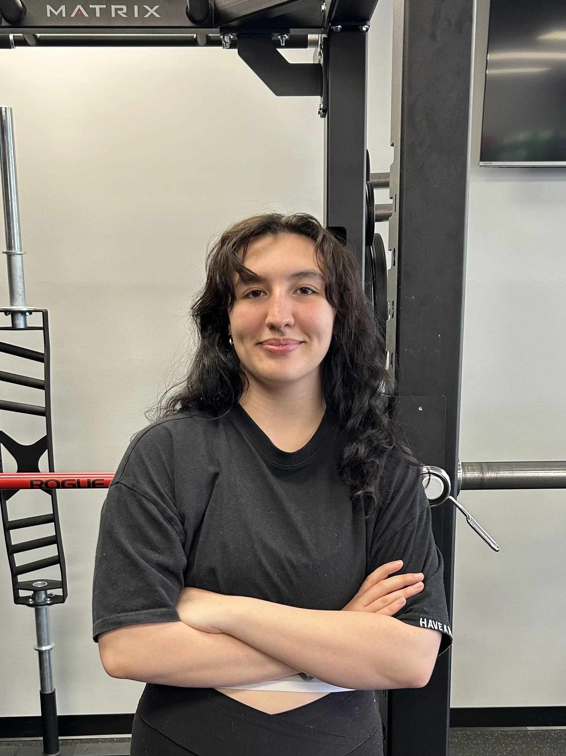 A young woman with curly black hair, wearing a black t-shirt and arms crossed, stands in a gym in front of fitness equipment.