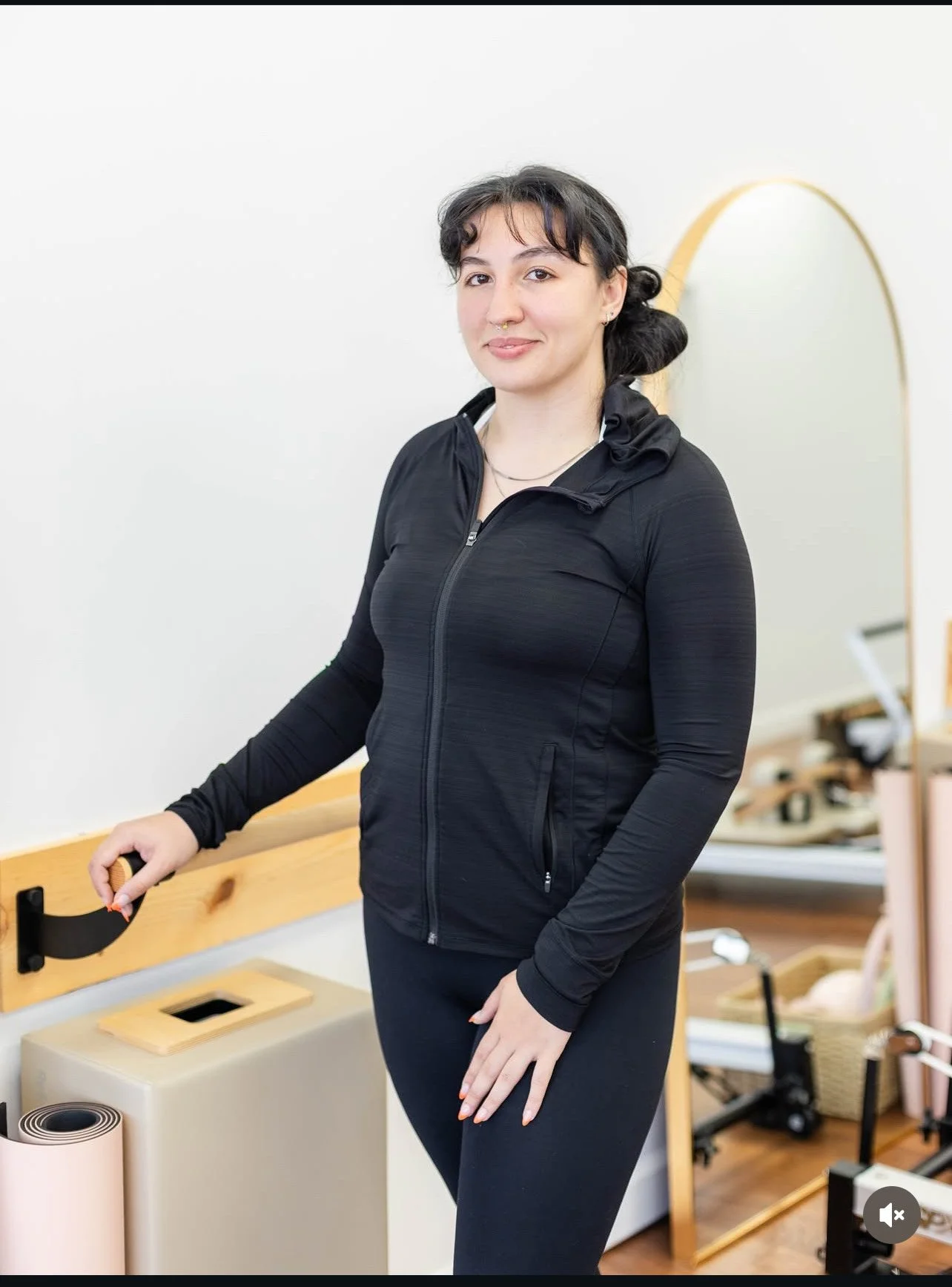A woman with dark hair, wearing a black athletic jacket and leggings, standing in a room with fitness equipment, holding a handle attached to a piece of exercise equipment.