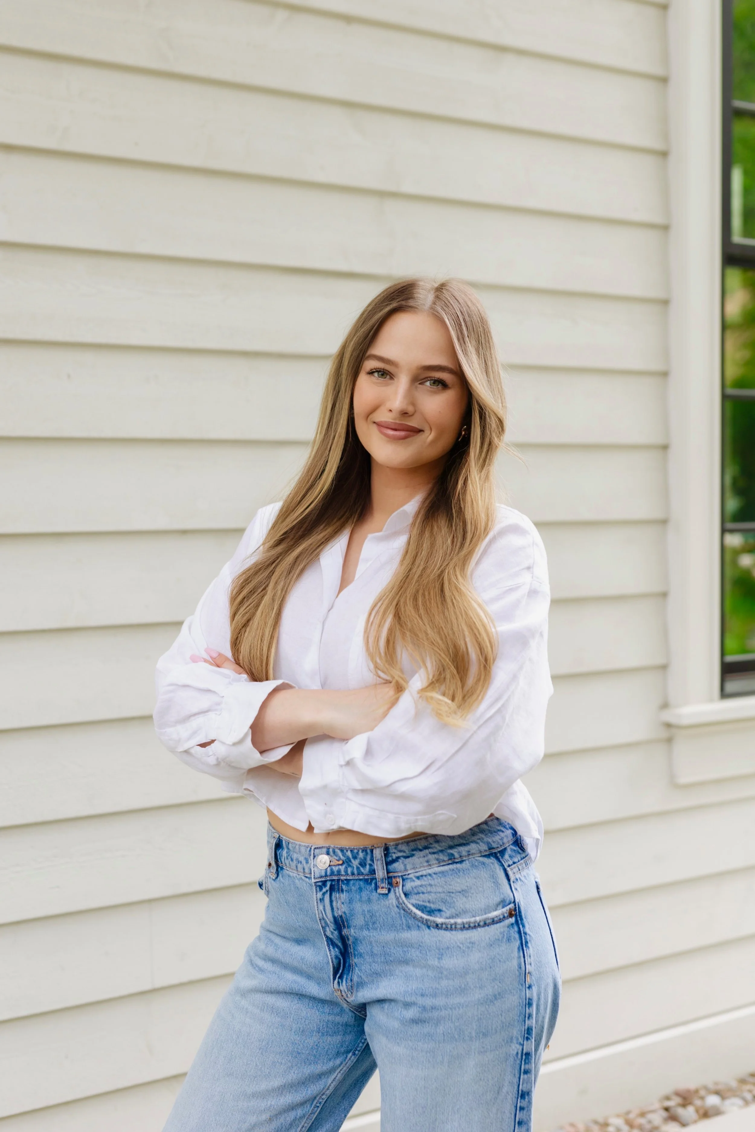Young woman with long wavy hair in a white shirt and blue jeans standing against a beige house exterior with arms crossed and smiling.