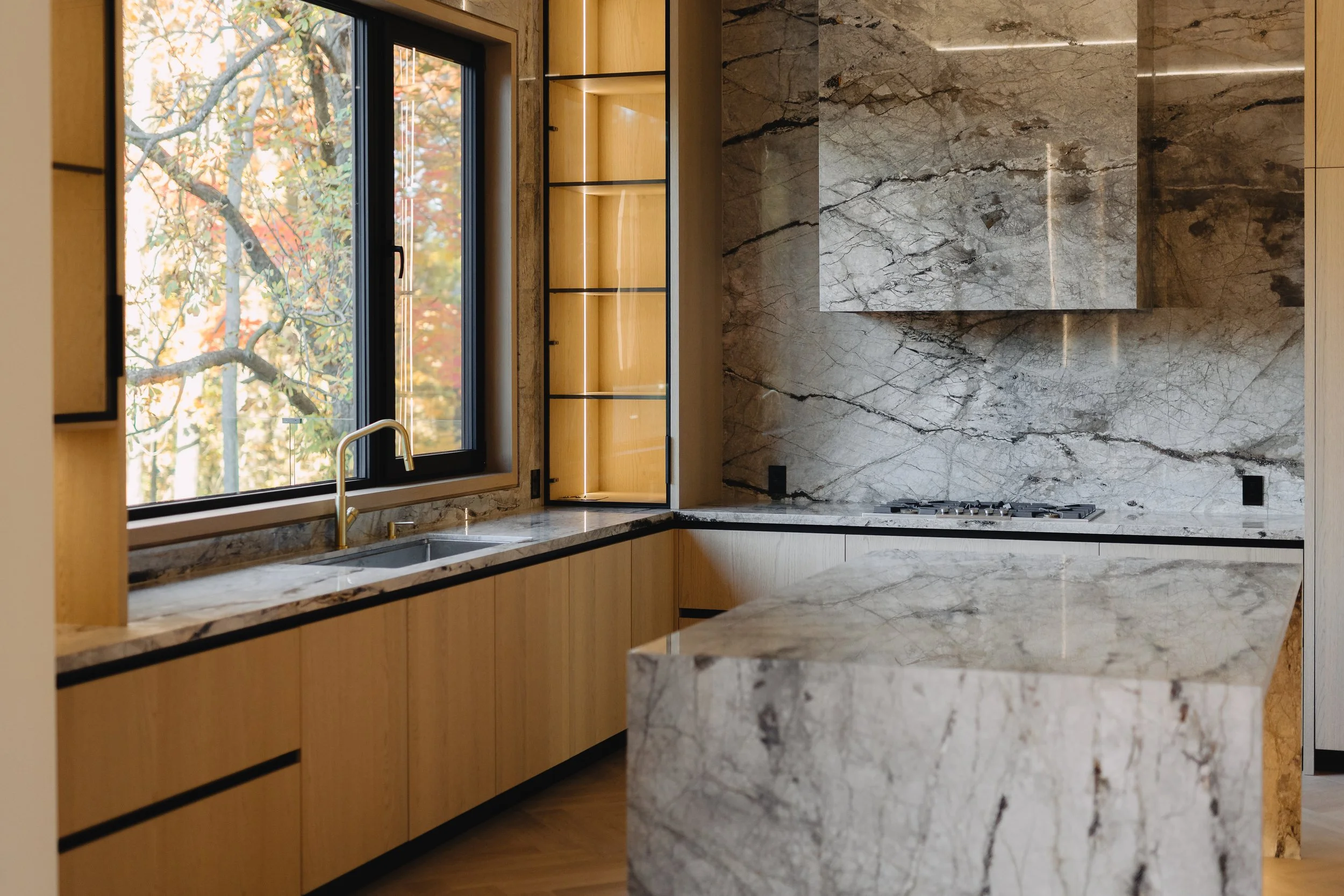 Modern kitchen with wooden cabinetry, marble countertops, and marble backsplash, featuring a large window with views of trees with autumn leaves.