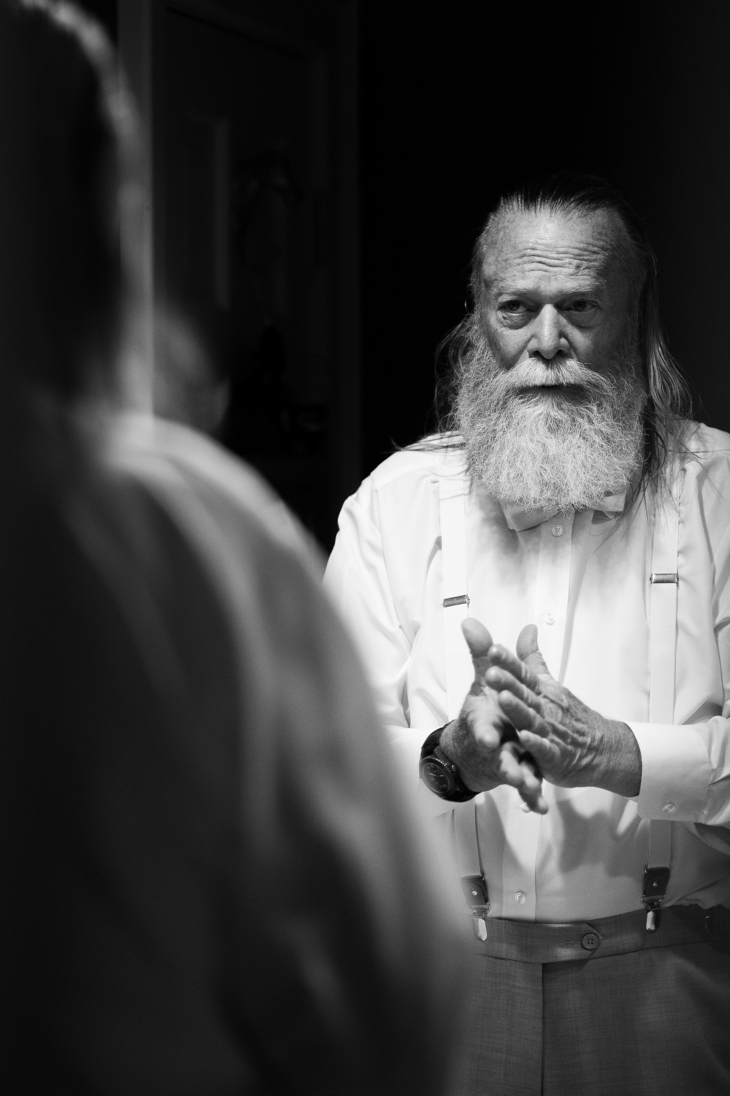 Black and white photo of an older man with a long white beard, dressed in a white shirt with suspenders, talking to someone whose back is turned to the camera.