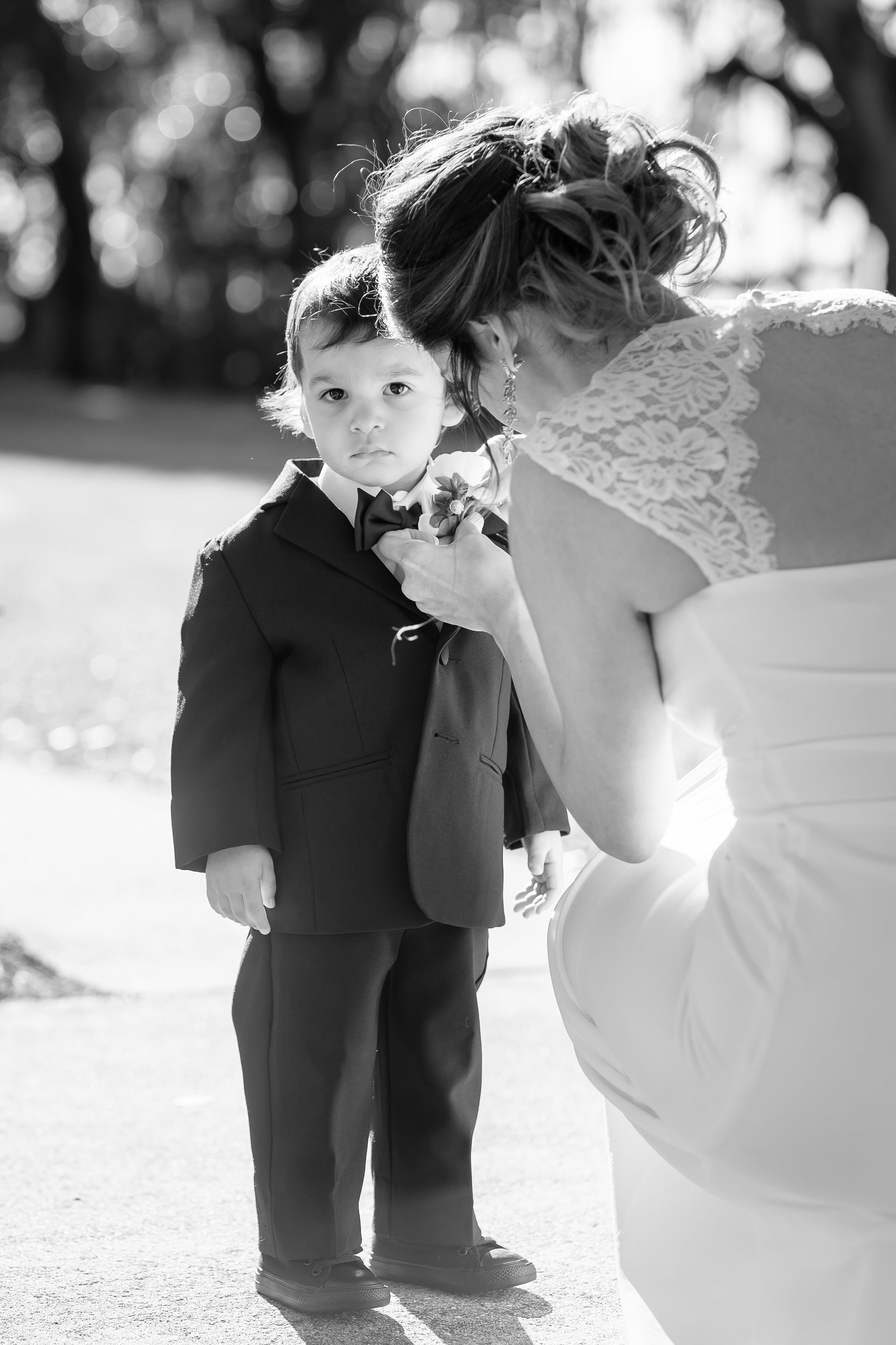 A young boy in a tuxedo stands outdoors as an adult woman arranges a boutonniere on his lapel. The woman is wearing a dress with lace details, and the background is blurred with trees and sunlight.