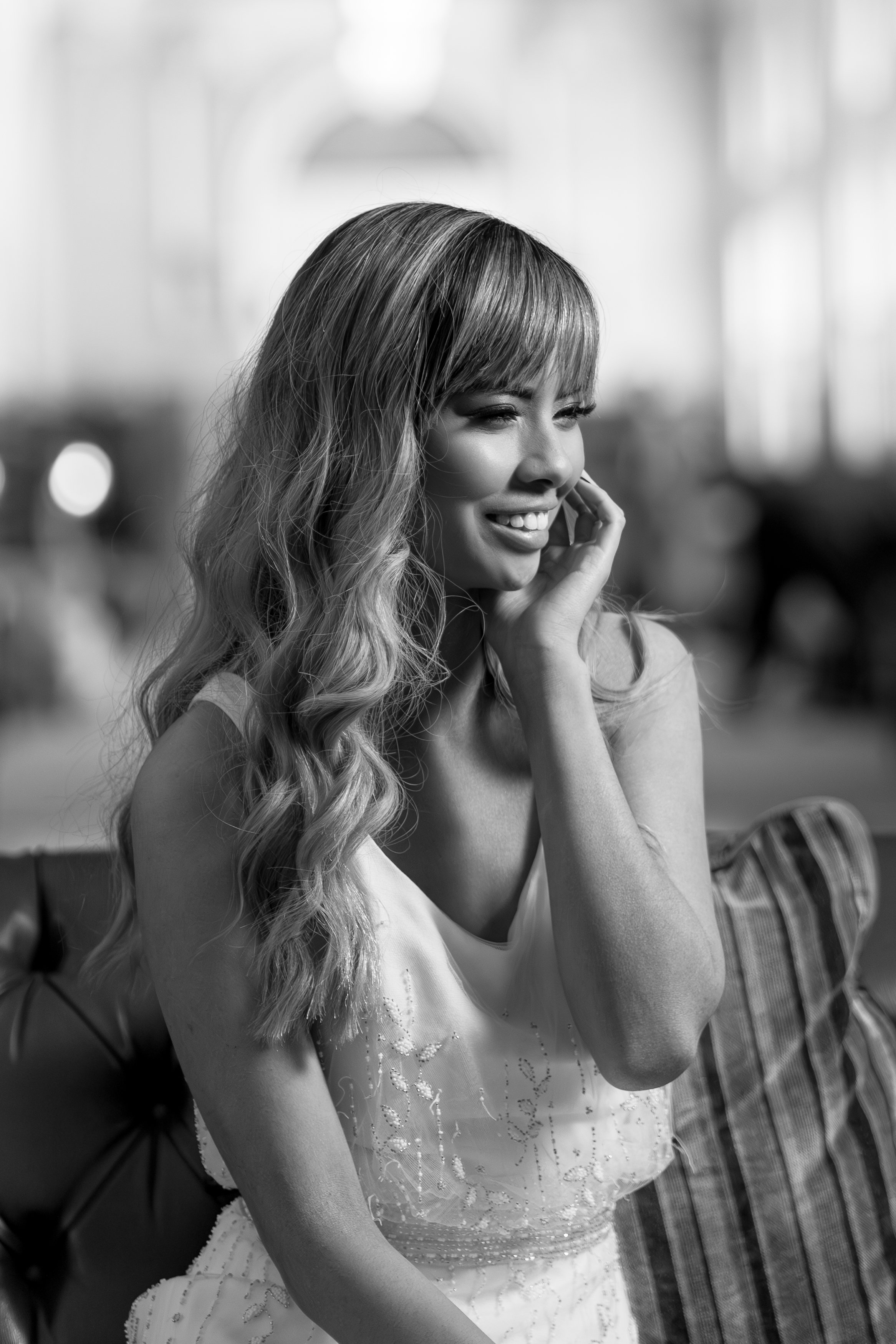 A black-and-white photo of a smiling woman with wavy hair, wearing a sleeveless dress, sitting on a bench indoors.