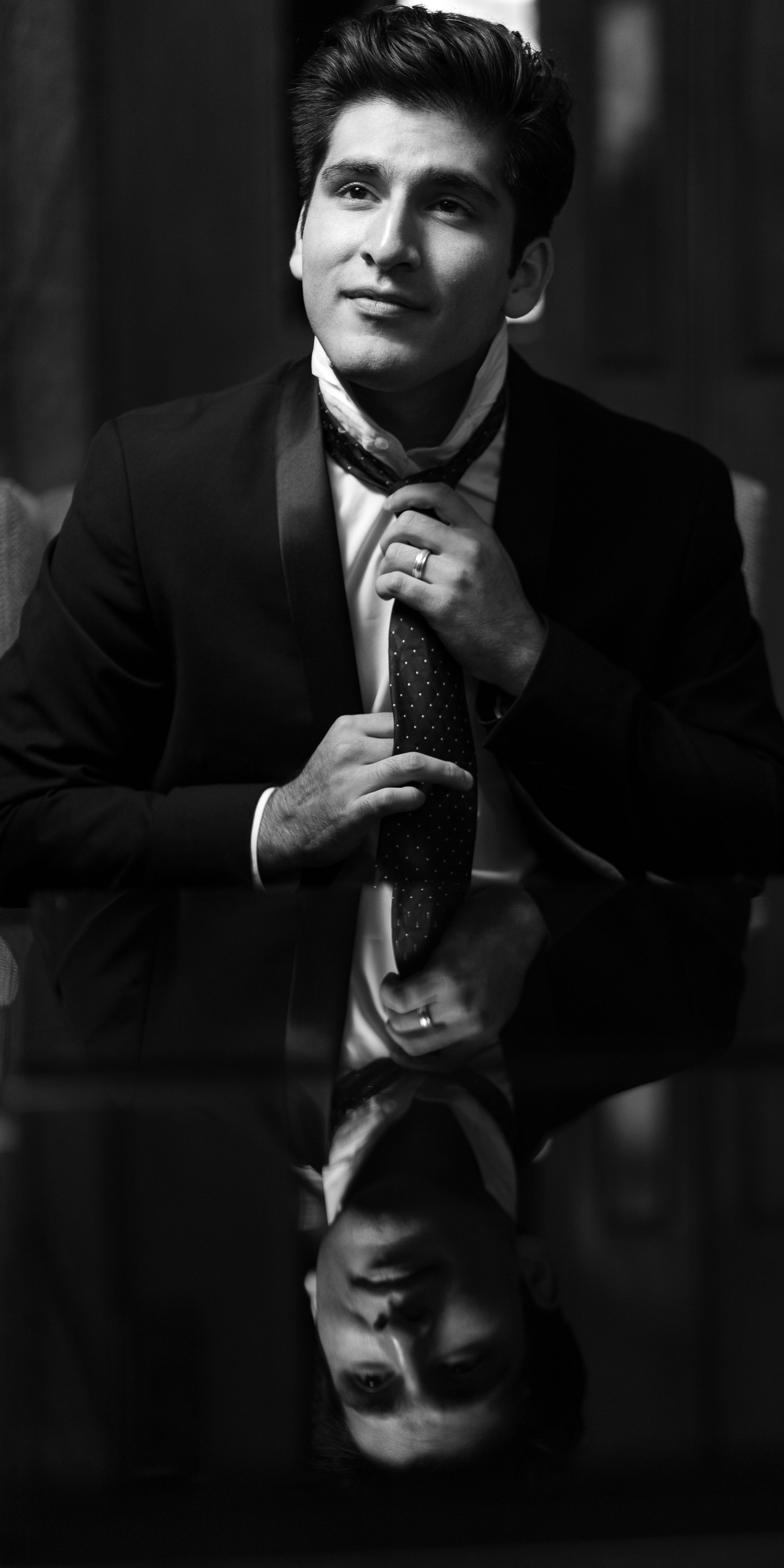 A young man in formal attire, adjusting a patterned tie, sitting at a reflective table, with his reflection visible beneath him in a black-and-white photograph.
