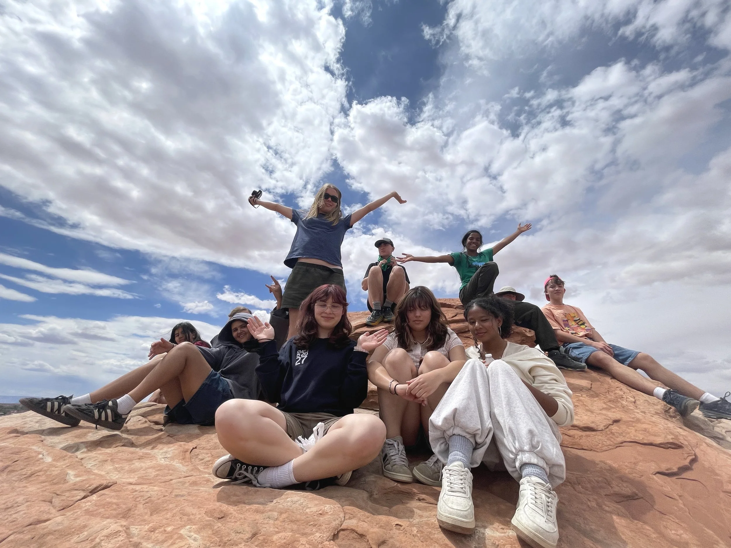 Group of nine students sitting and standing on red rock formation outdoors under partly cloudy sky.