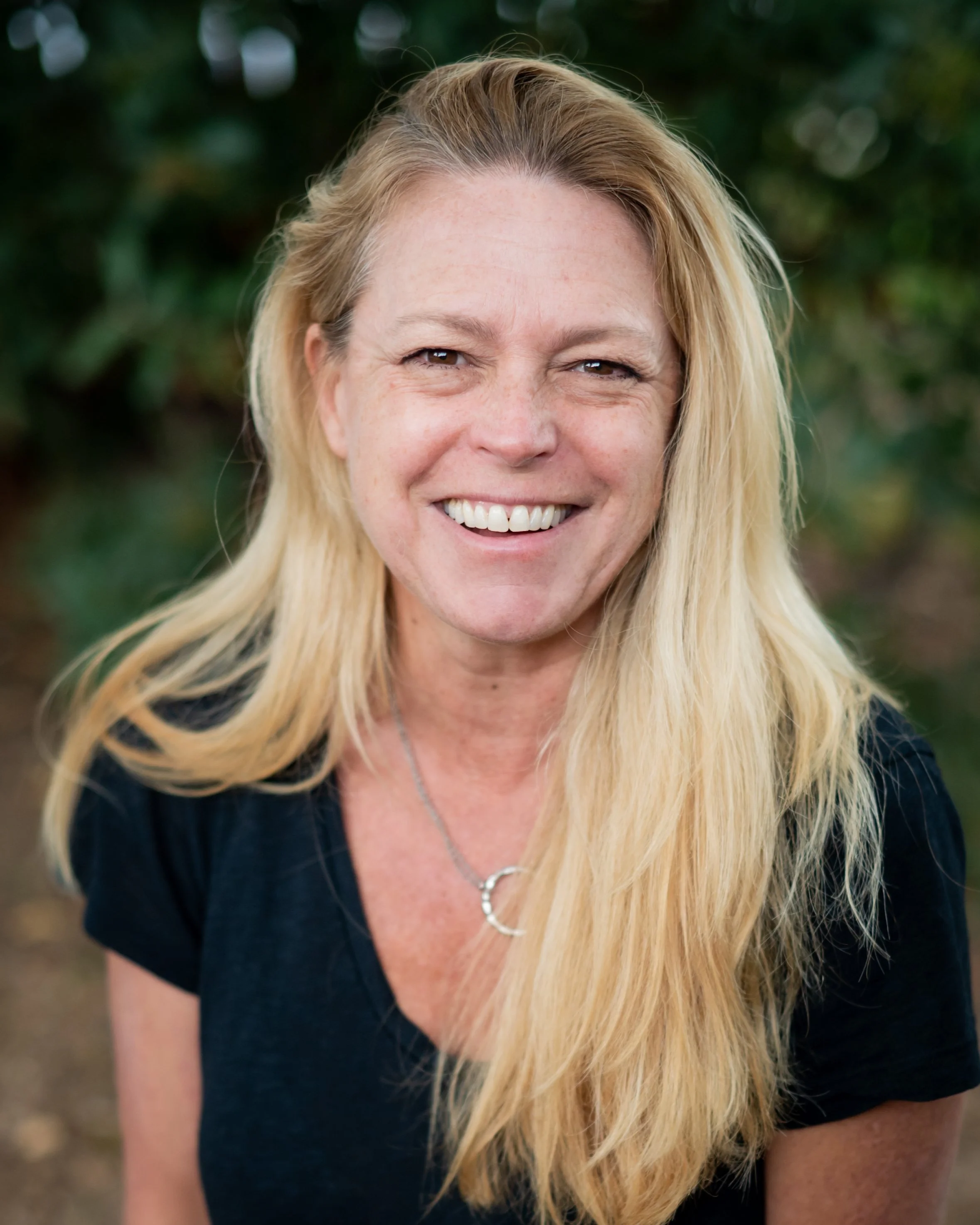 A woman with long blonde hair, wearing a pink shirt and a turquoise necklace, smiling outdoors.