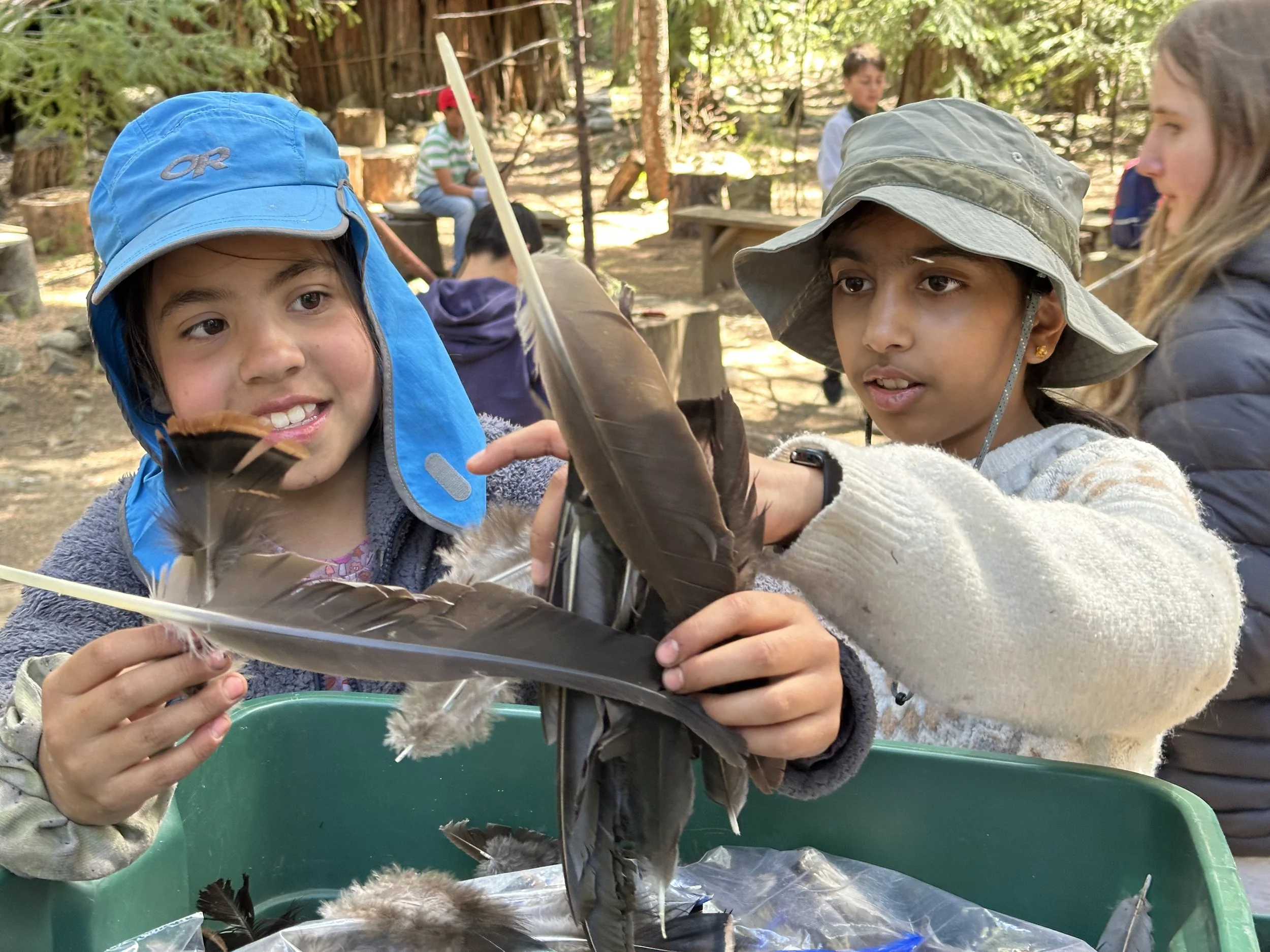 Two young female students wearing hats and jackets examining feathers and possibly small animal pelts outdoors.