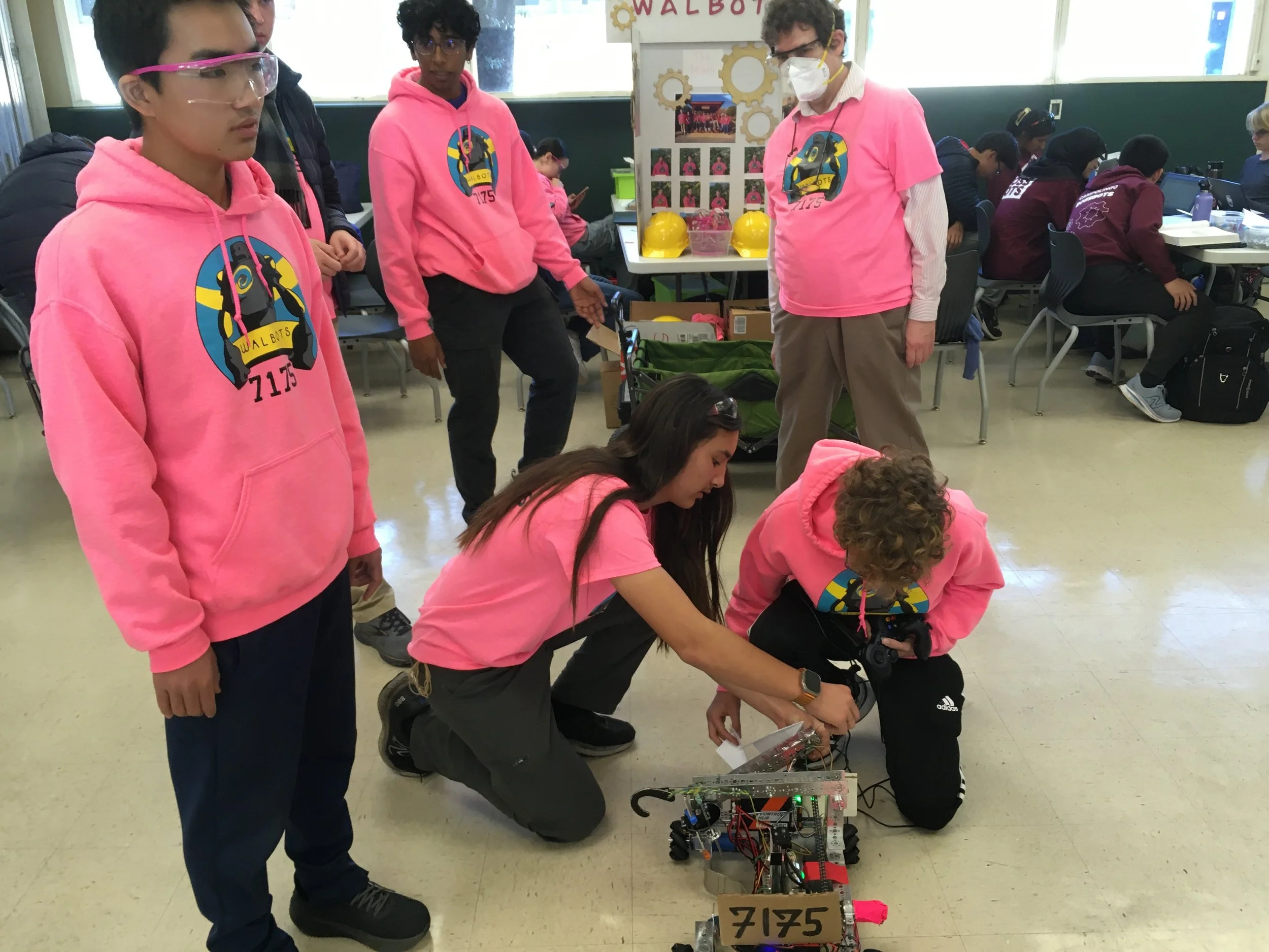 Group of students and teachers wearing pink hoodies working on a robot, in a classroom with other students in the background.