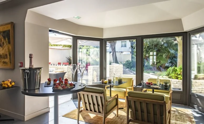 Salon lumineux avec grande baie vitrée, fauteuils en bois, table basse, plantes vertes, et décoration moderne.