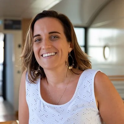 Femme souriante avec des boucles d'oreilles longues, portant un haut blanc à trous, dans un intérieur moderne.