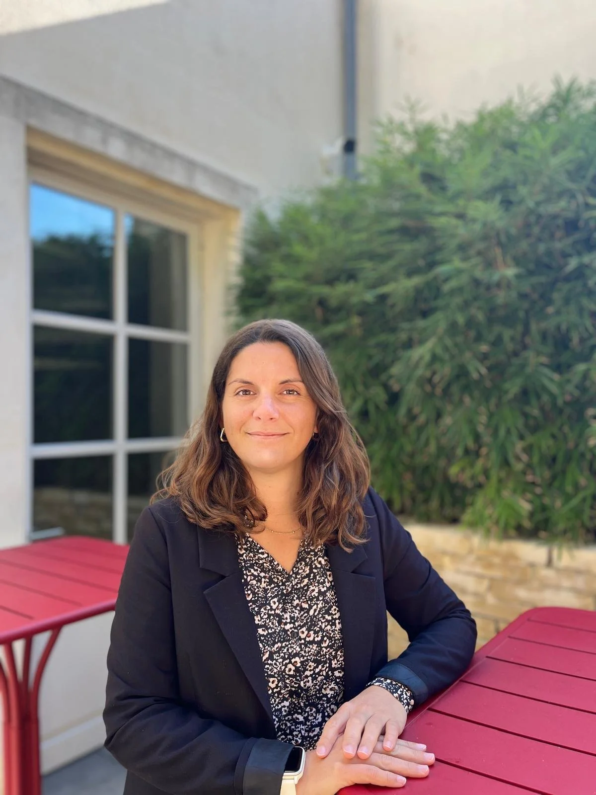 Une femme souriante avec des cheveux bruns, portant un blazer noir et une robe à motifs, assise à une table rouge à l'extérieur, avec un grand arbre et une maison en arrière-plan.