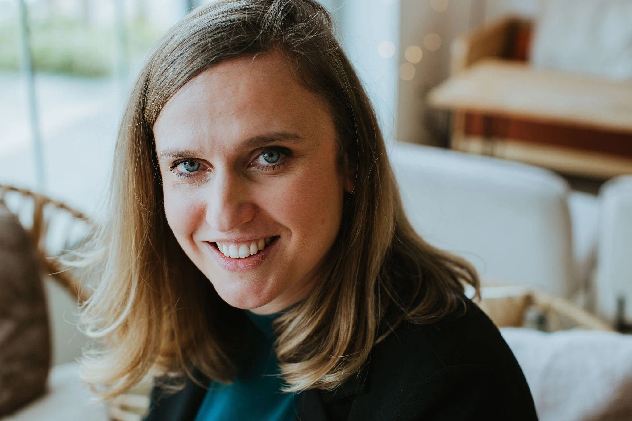 Une femme aux cheveux châtains, aux yeux bleus, souriante, dans un intérieur lumineux avec un canapé blanc en arrière-plan.