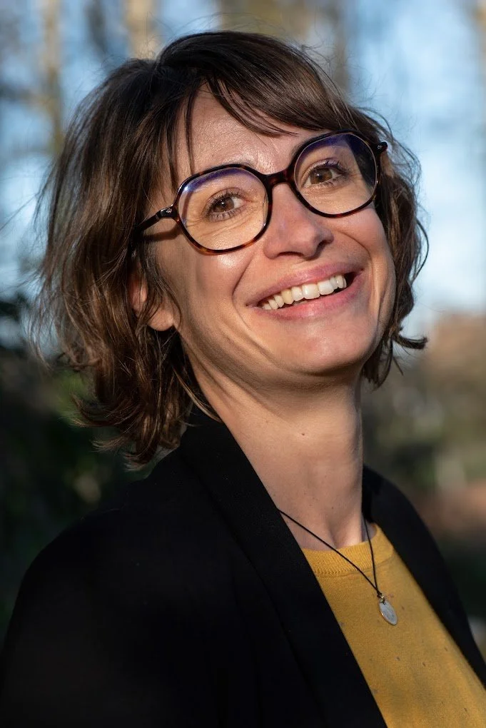 Une femme souriante portant des lunettes rondes, avec des cheveux courts et bouclés, portant un pull jaune, une veste noire et une chaîne en collier, en extérieur.