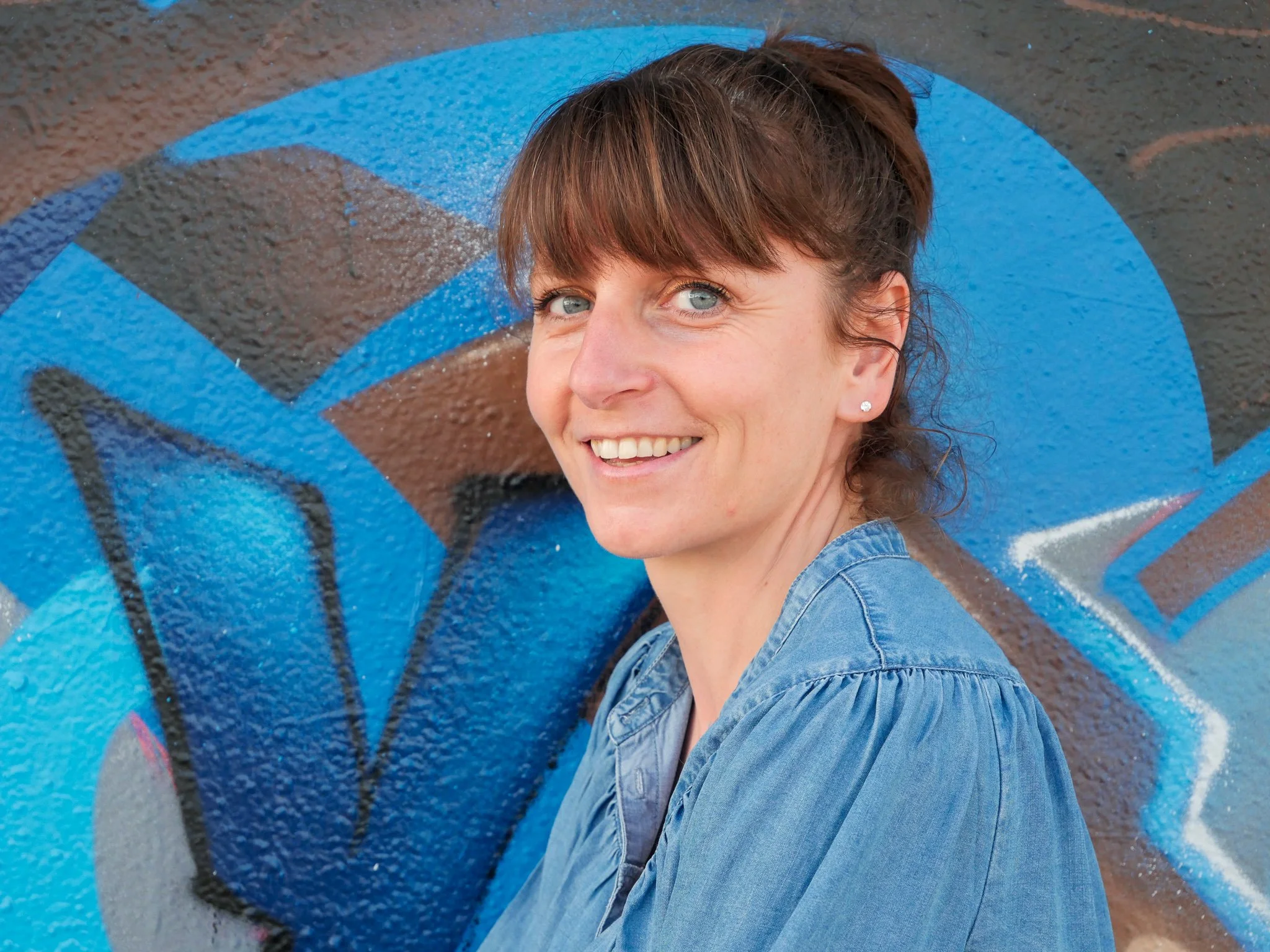 Femme souriante avec des cheveux bruns et des yeux bleus, portant une veste en jean, posant devant un mur avec graffitis bleus, noirs et grays.