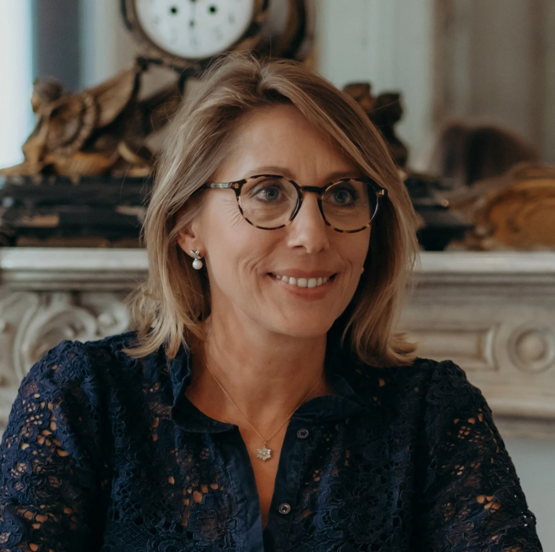 Une femme souriante avec des lunettes écaille, portant des boucles d'oreilles perles et un collier, assise dans une pièce décorée avec une cheminée en bois et une horloge ancienne en arrière-plan.