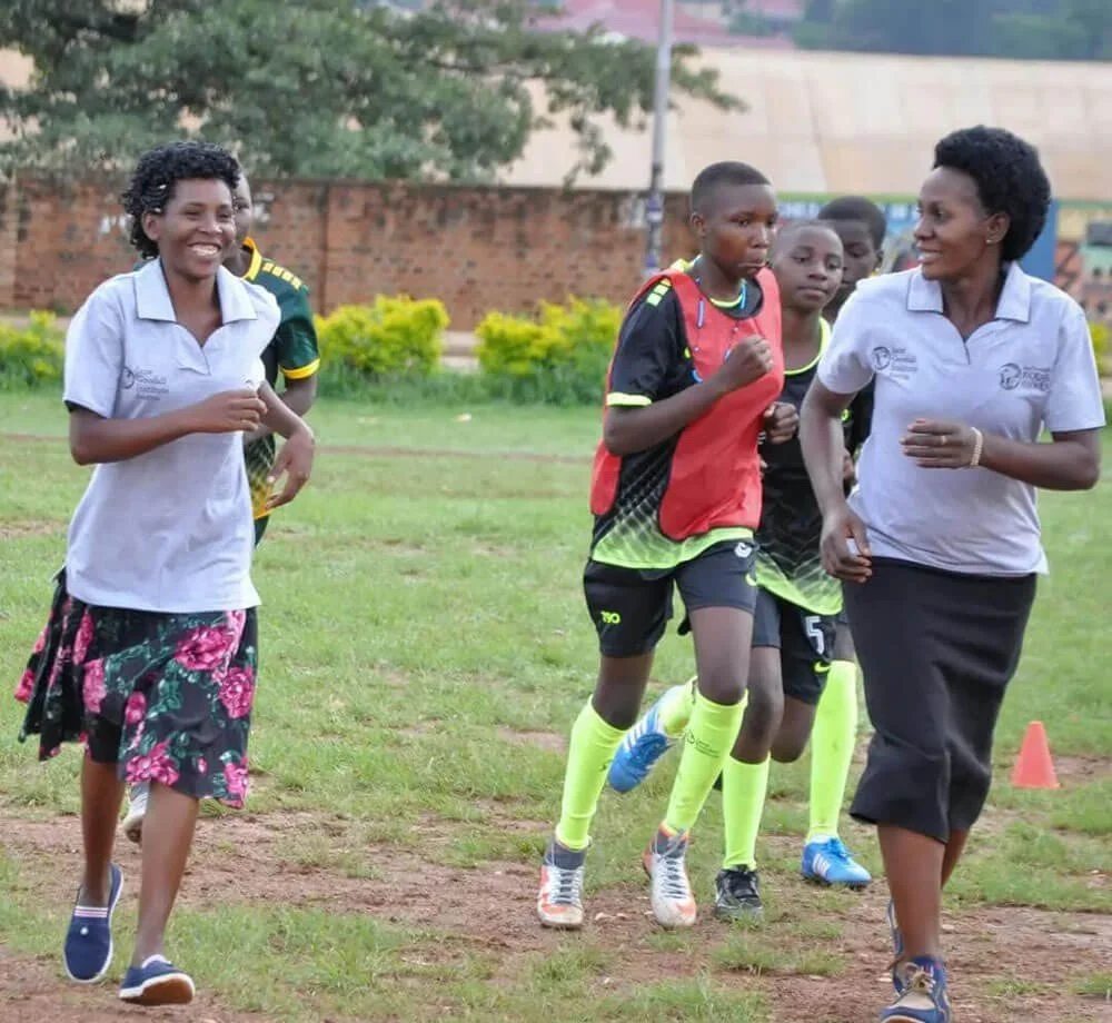 Young athletes running on a track field with two women, possibly coaches or teachers, encouraging them, outdoors on a grassy field with trees and a brick wall in the background.