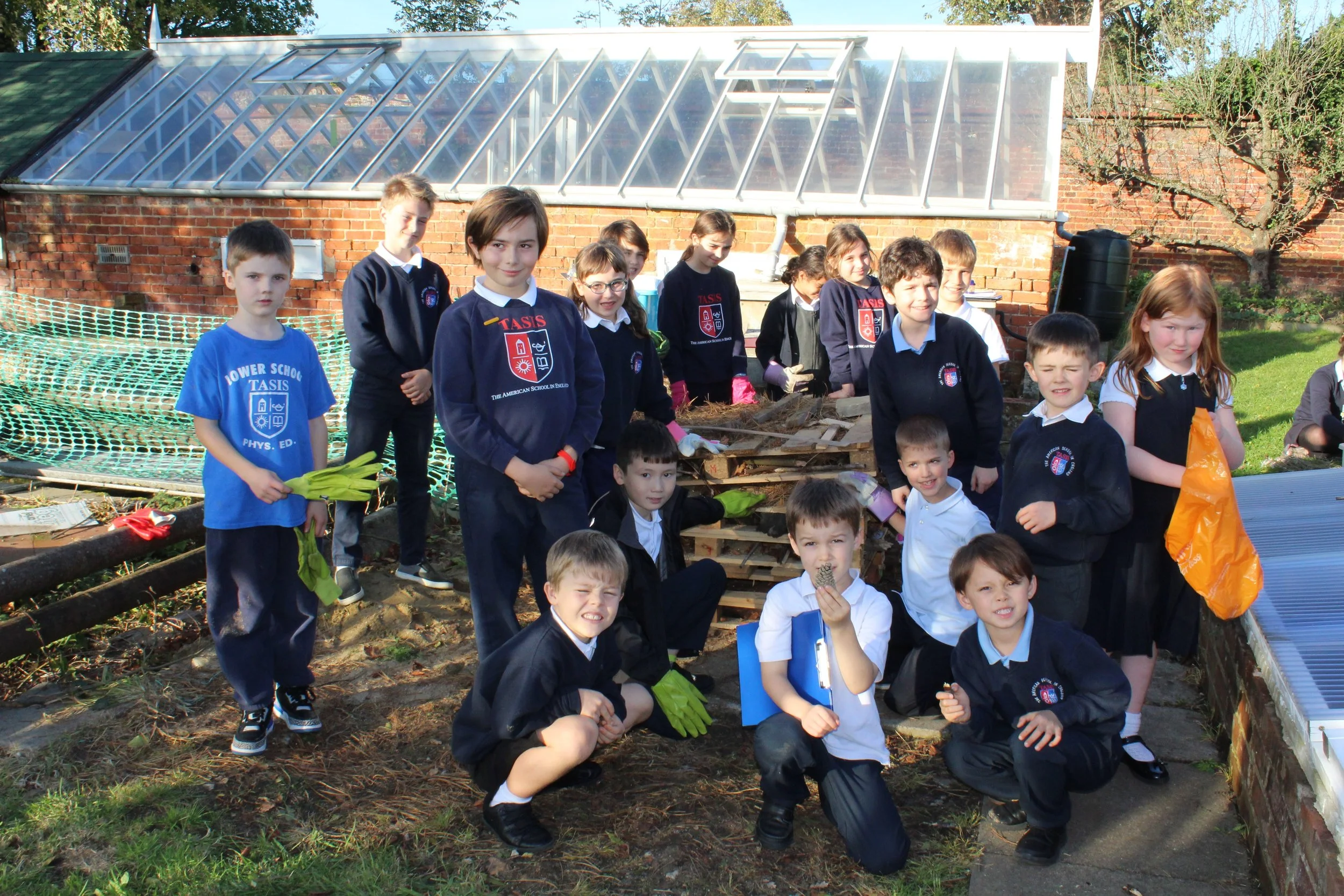 Group of elementary school children in school uniforms working on a gardening project outside on a sunny day, some holding tools and gloves. They are gathered around a pile of wood and soil near greenhouses and a brick wall.