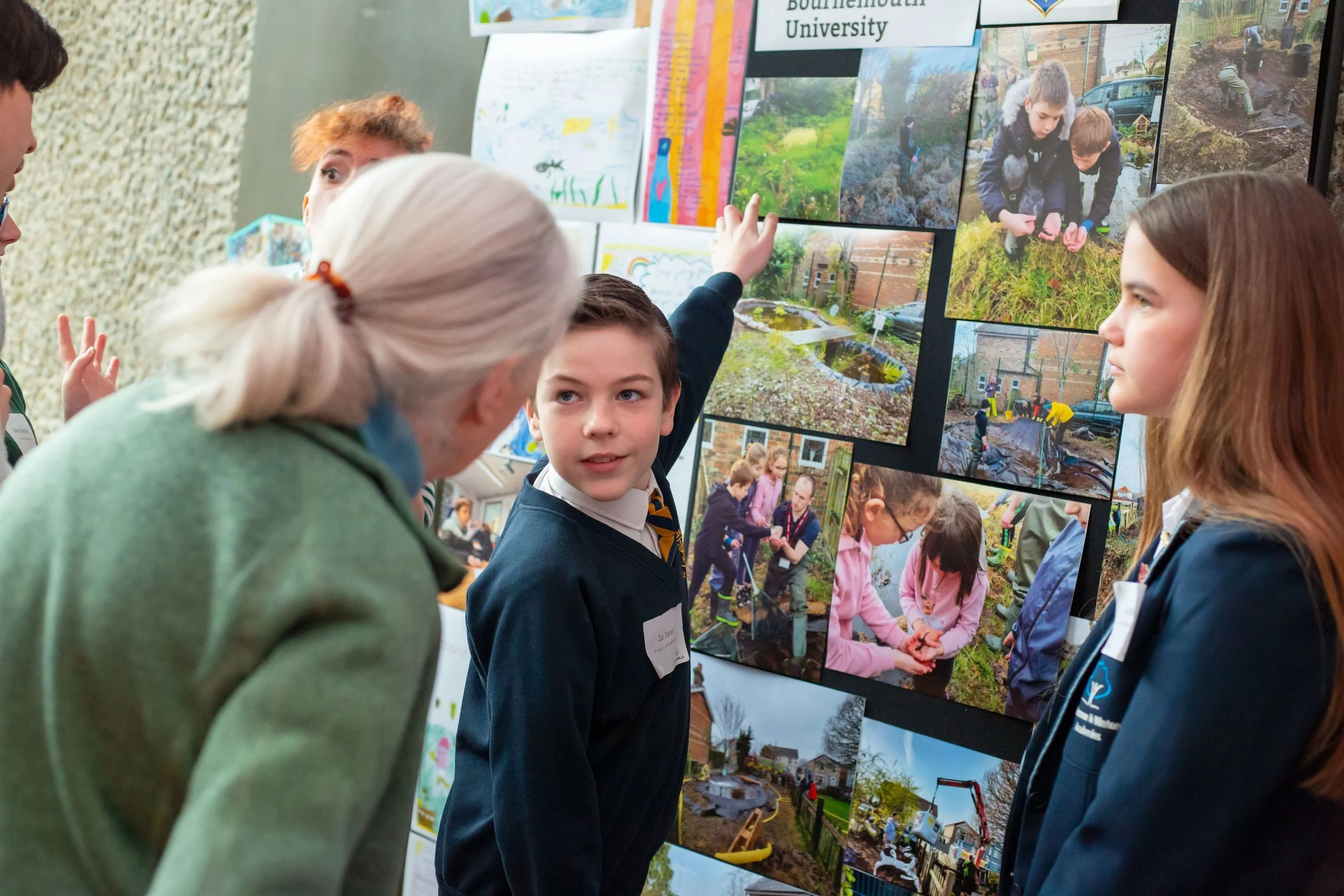 Students and a teacher discussing photographs of outdoor projects displayed on a board at an event.