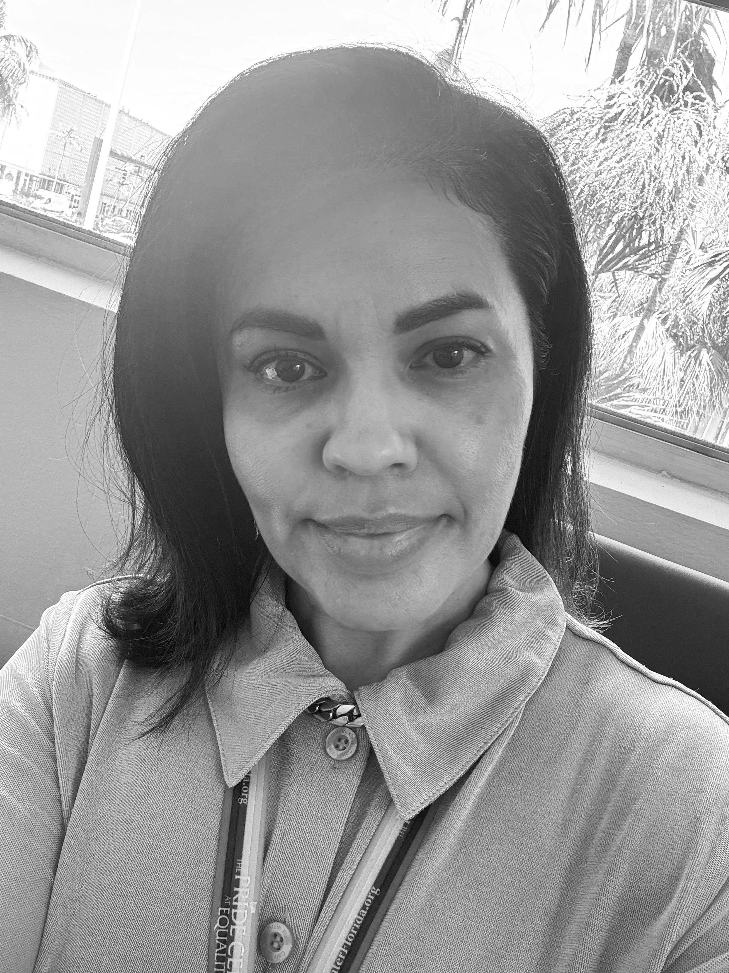 A woman with dark hair and a light-colored collared shirt taking a selfie indoors near a window with a cityscape and palm trees outside.