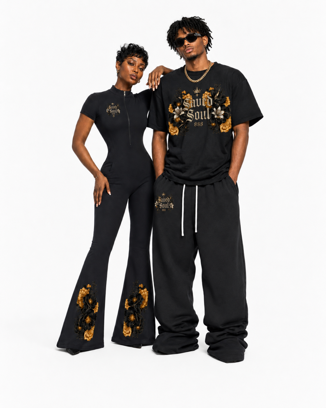 Two models standing against a white background, wearing black clothing with gold and black floral designs and the words "Saved Soul". The woman has short curly black hair, sunglasses, and wears a fitted jumpsuit, while the man has medium-length curly