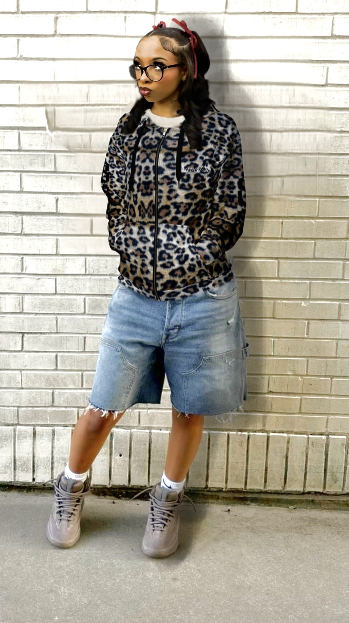 Young woman standing against a white brick wall, dressed in a leopard print jacket, baggy denim shorts, and athletic shoes, with glasses and styled hair with red ribbons.